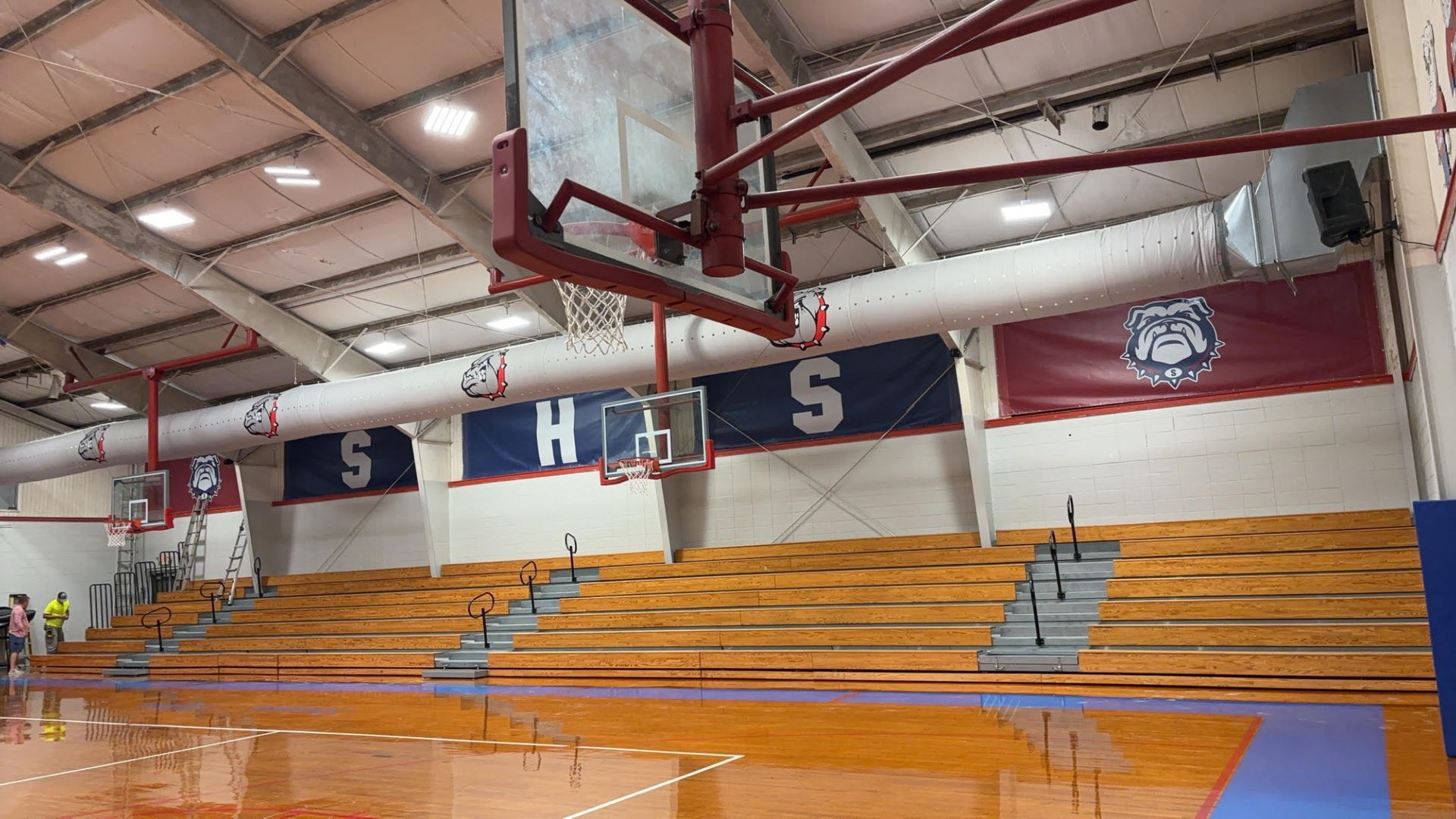 A high-school basketball gym featuring wooden bleachers, a basketball hoop, and banners displaying 