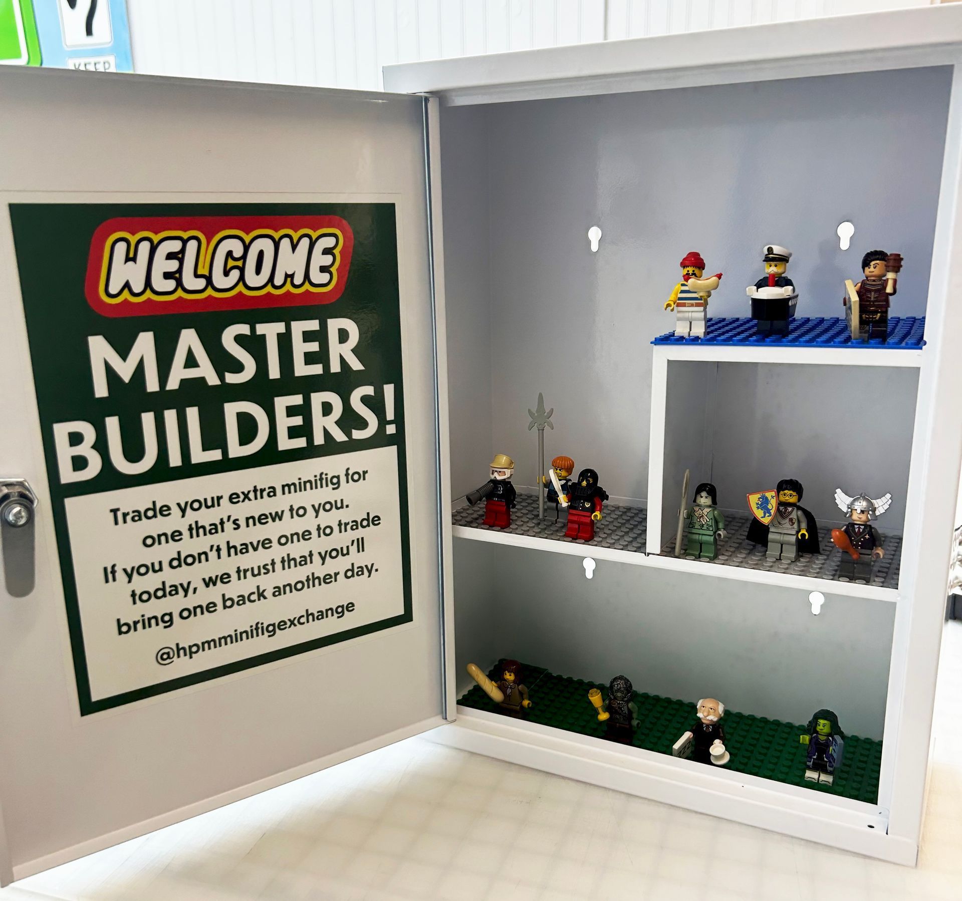 A wall-mounted display case containing LEGO minifigures for trade, featuring a sign inviting visitors to exchange figures.