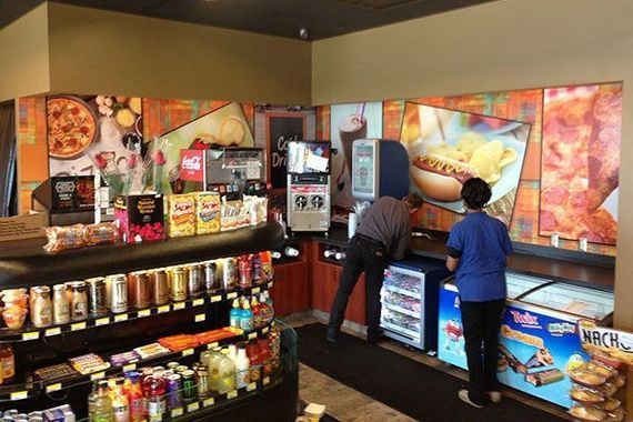 Inside a convenience store with food displays and drink machines, two staff members work behind the service counter.