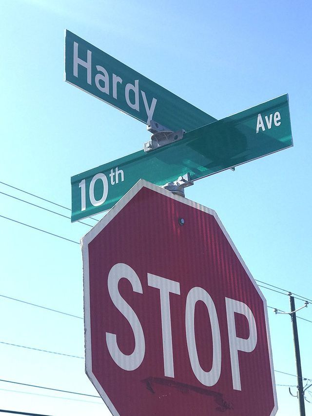 A stop sign sits in front of green street signs for Hardy and 10th Ave against a blue sky.