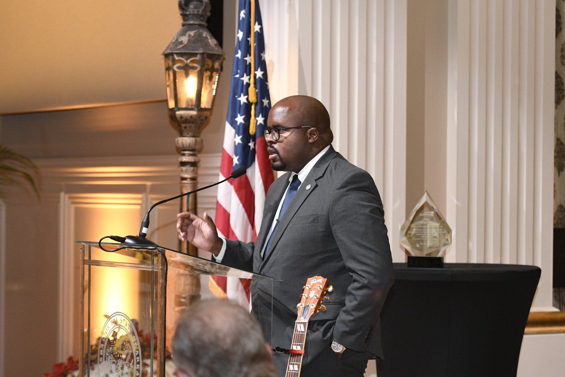 A man in a suit is giving a speech at a podium in front of an american flag.