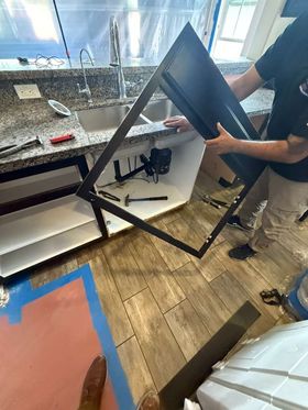 A person installing a black cabinet door frame beneath a granite kitchen sink.