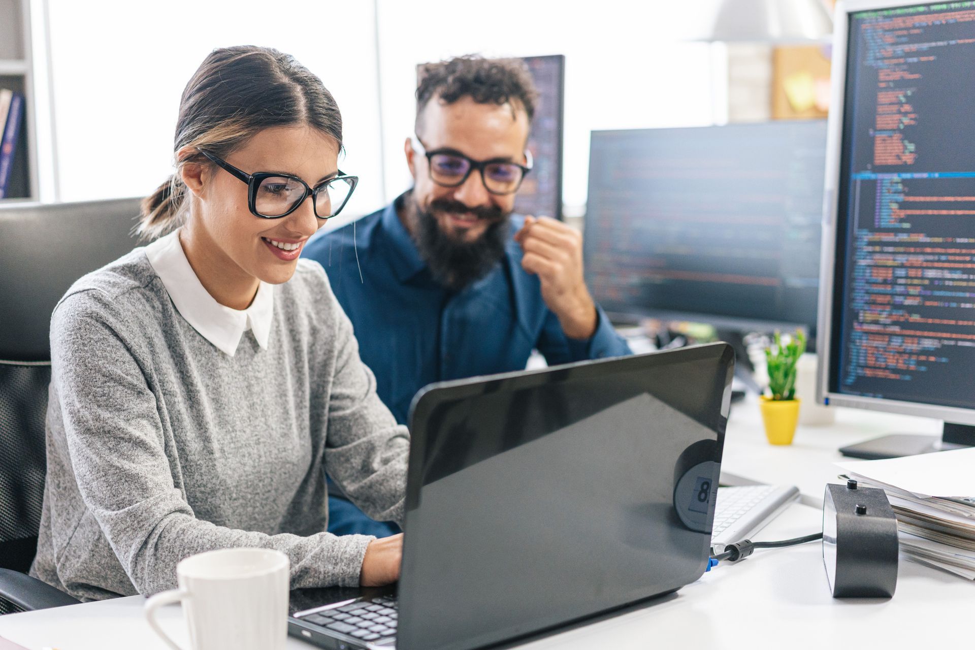 A man and a woman are sitting at a desk looking at a laptop computer.