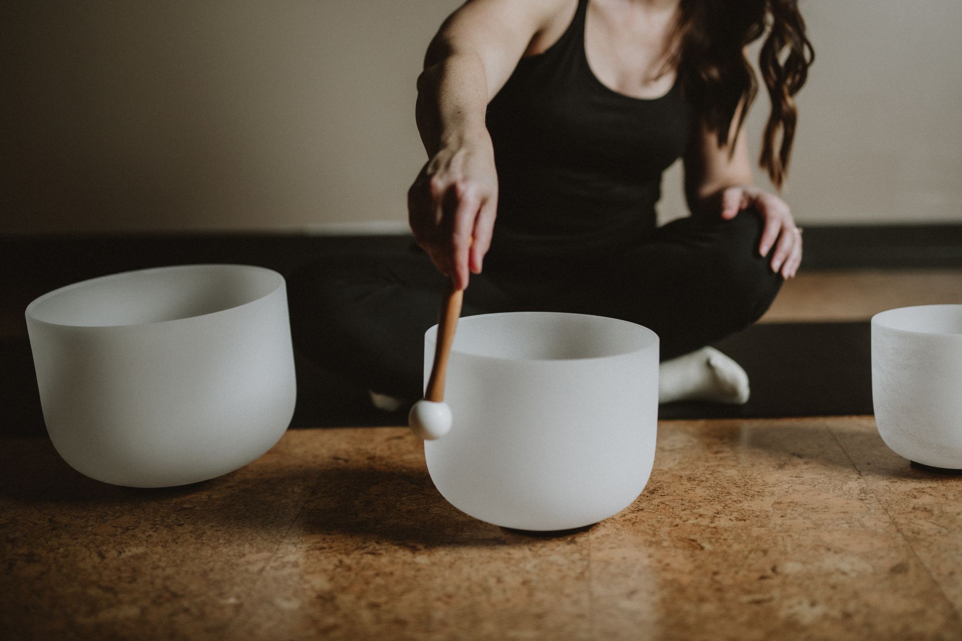 A person sits on a cork floor, gently striking a white quartz singing bowl with a wooden mallet during a sound bath.