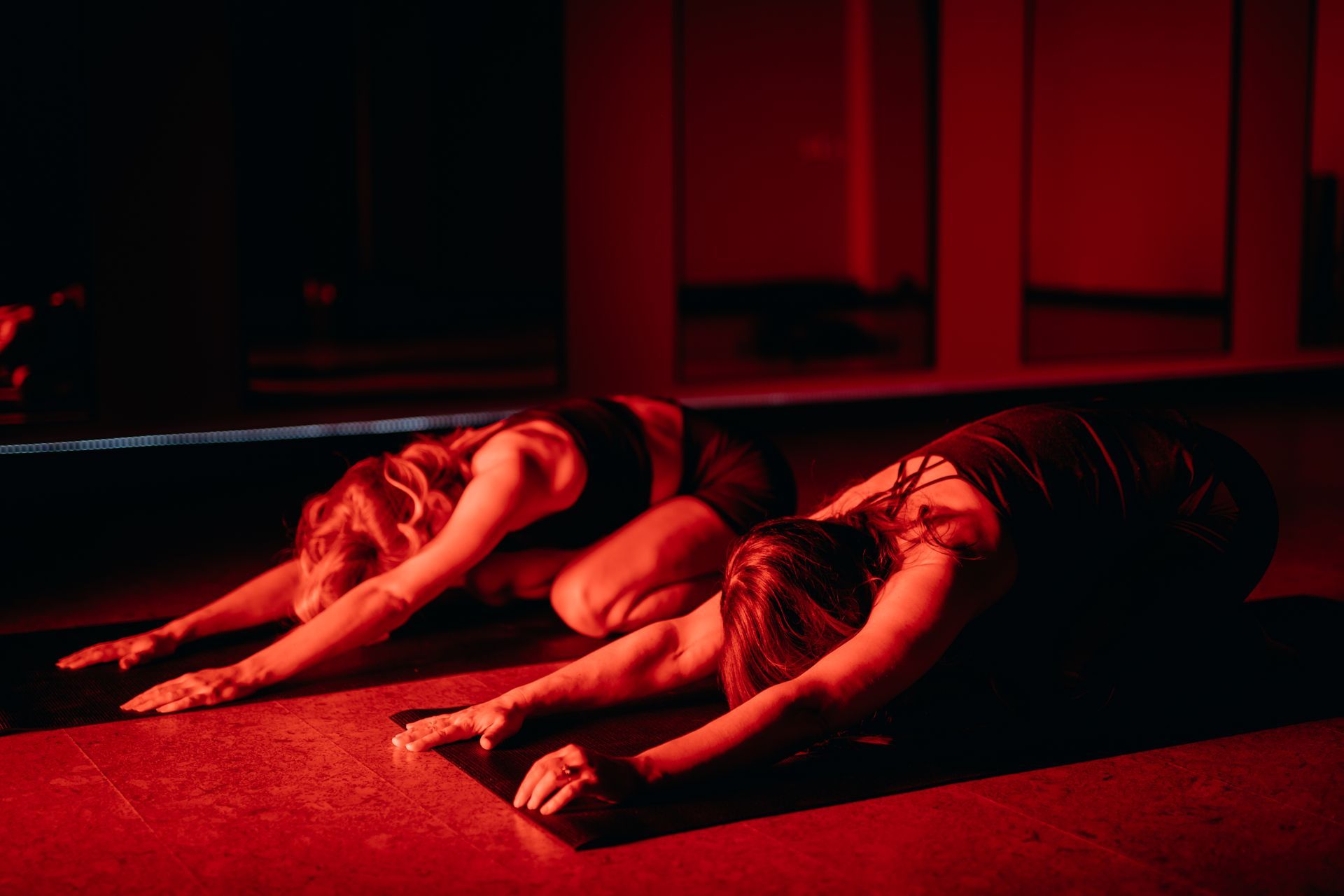 Two people in black workout attire practice child's pose on yoga mats in a dark room illuminated by deep red light.
