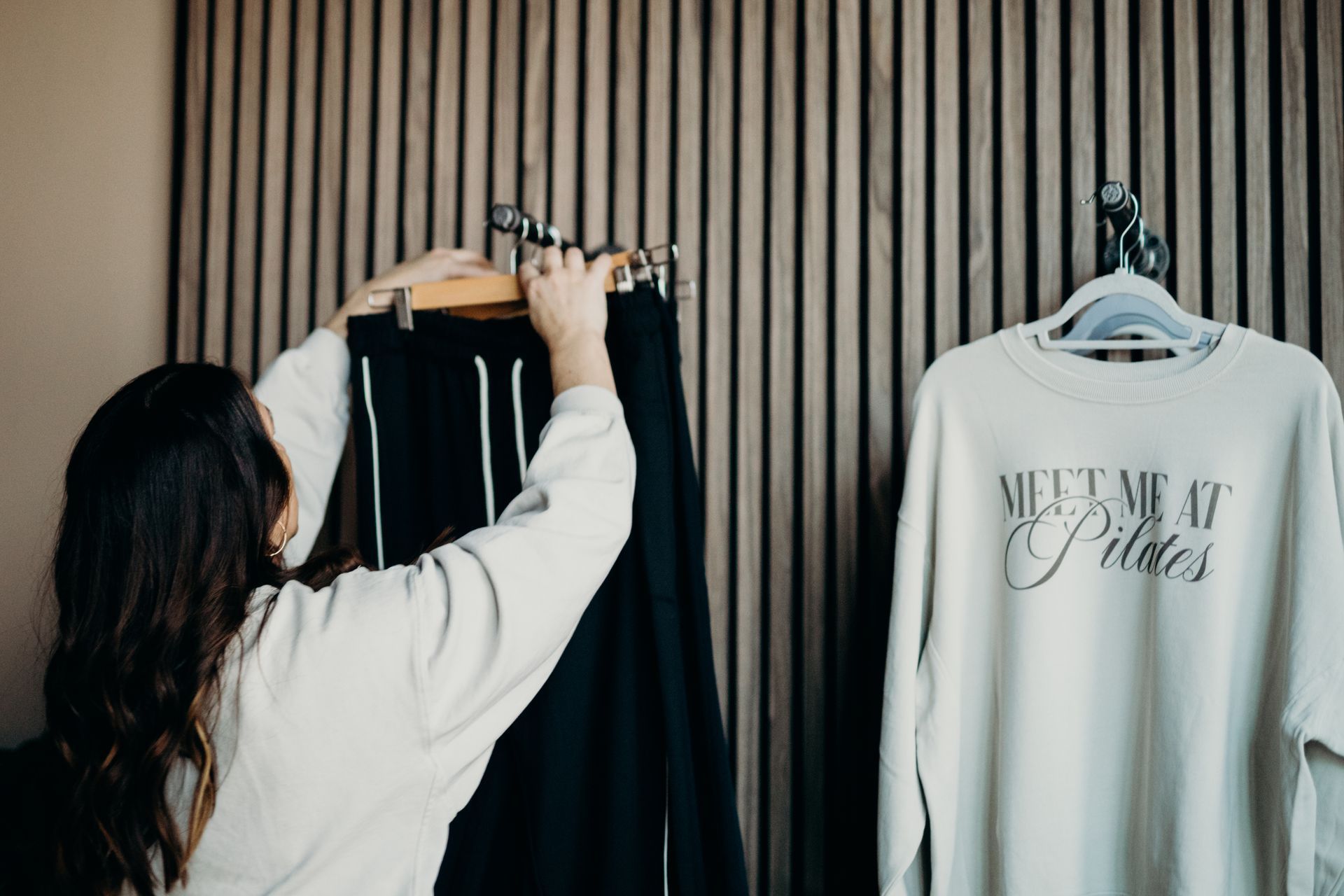 A person in a cream sweatshirt hangs black pants on a rack next to a sweatshirt printed with 