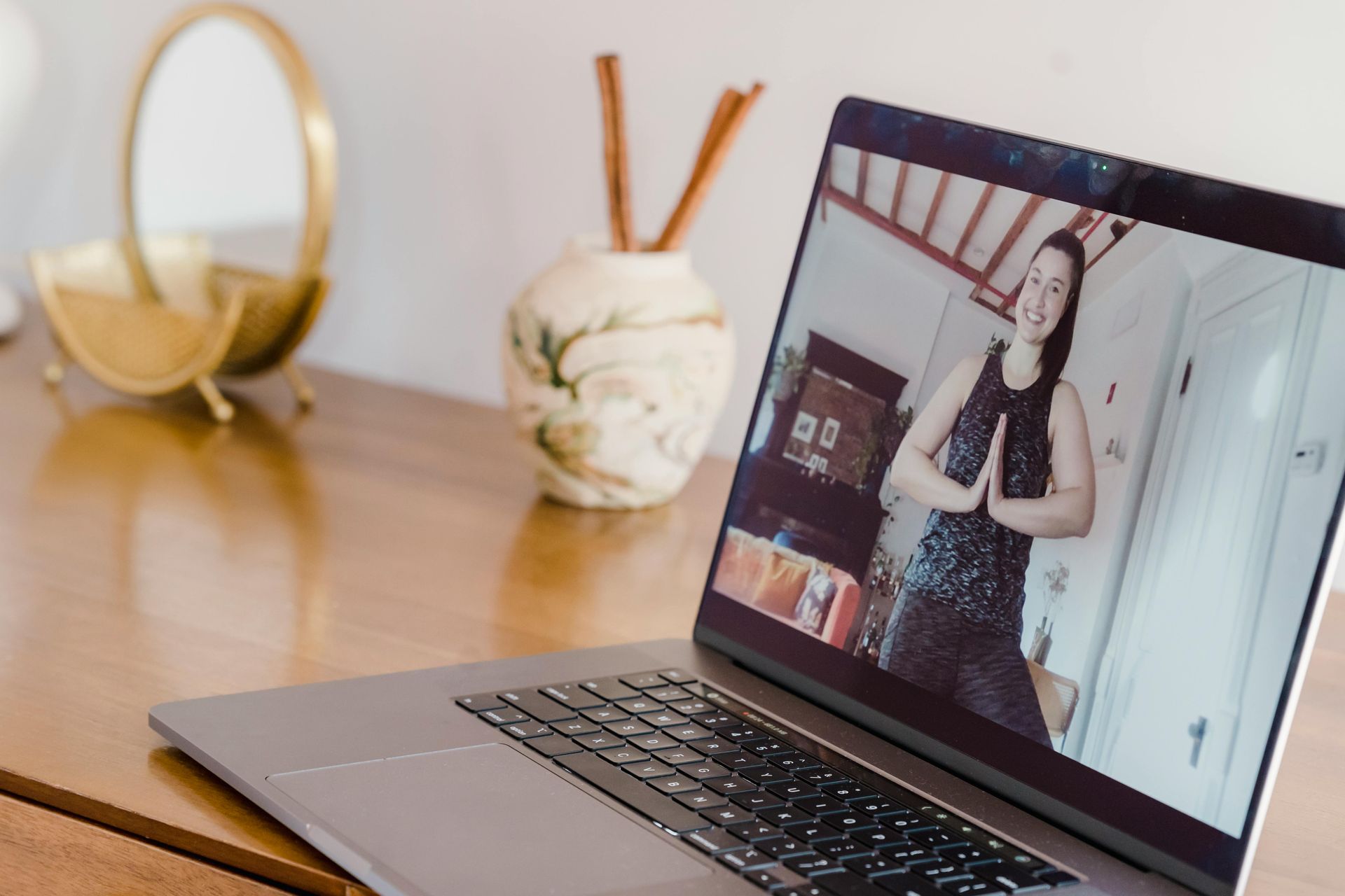 A laptop displaying a virtual fitness instructor in a prayer pose, sitting on a wooden desk with a mirror and vase.