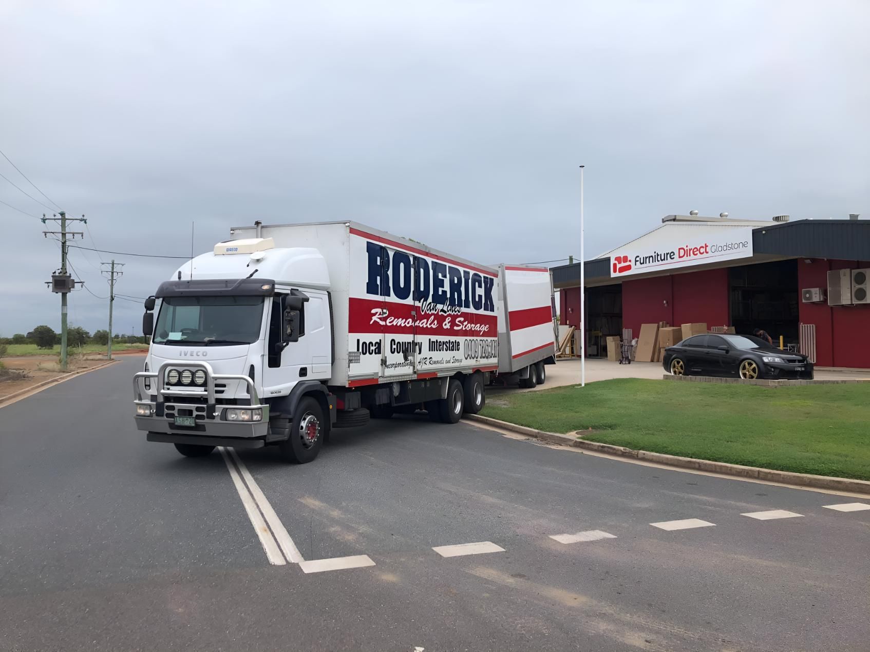 A Red and White Truck is Driving Down a Road Next to a Building — Roderick Removals & Van Lines In Gold Coast, NSW