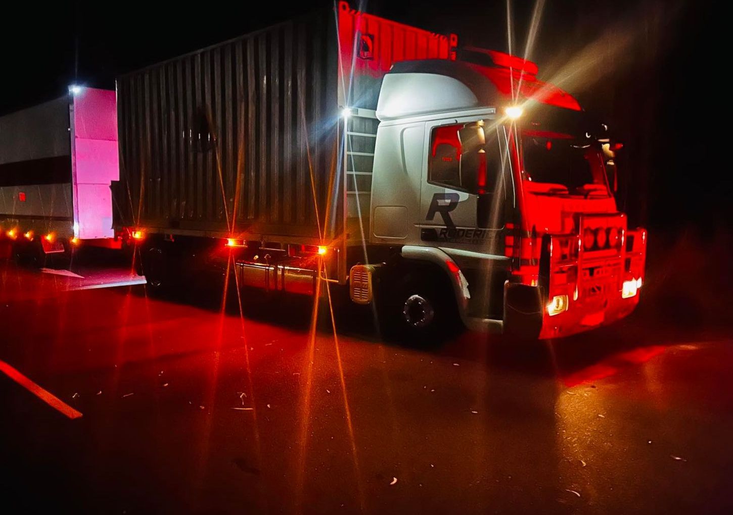 A large white semi-truck with two container trailers is parked on a wet road at night.