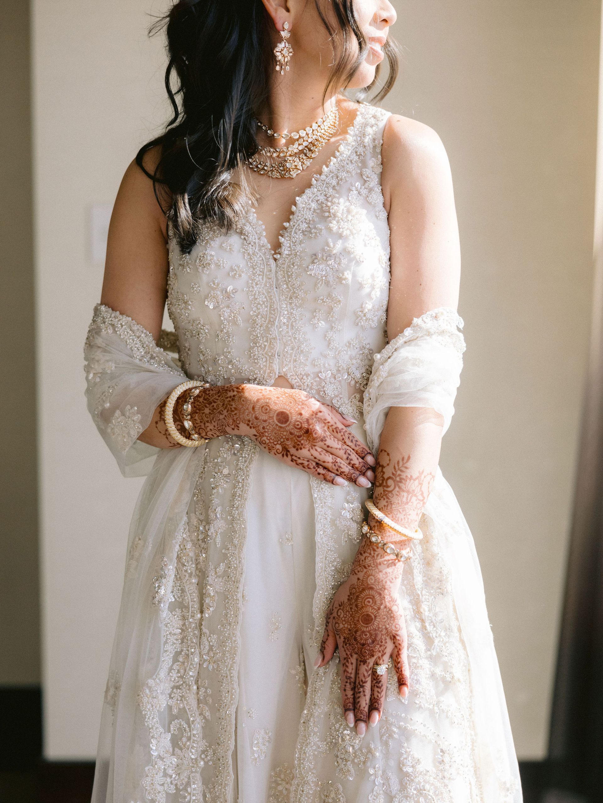 A woman in a white dress with henna on her hands is standing in front of a window.