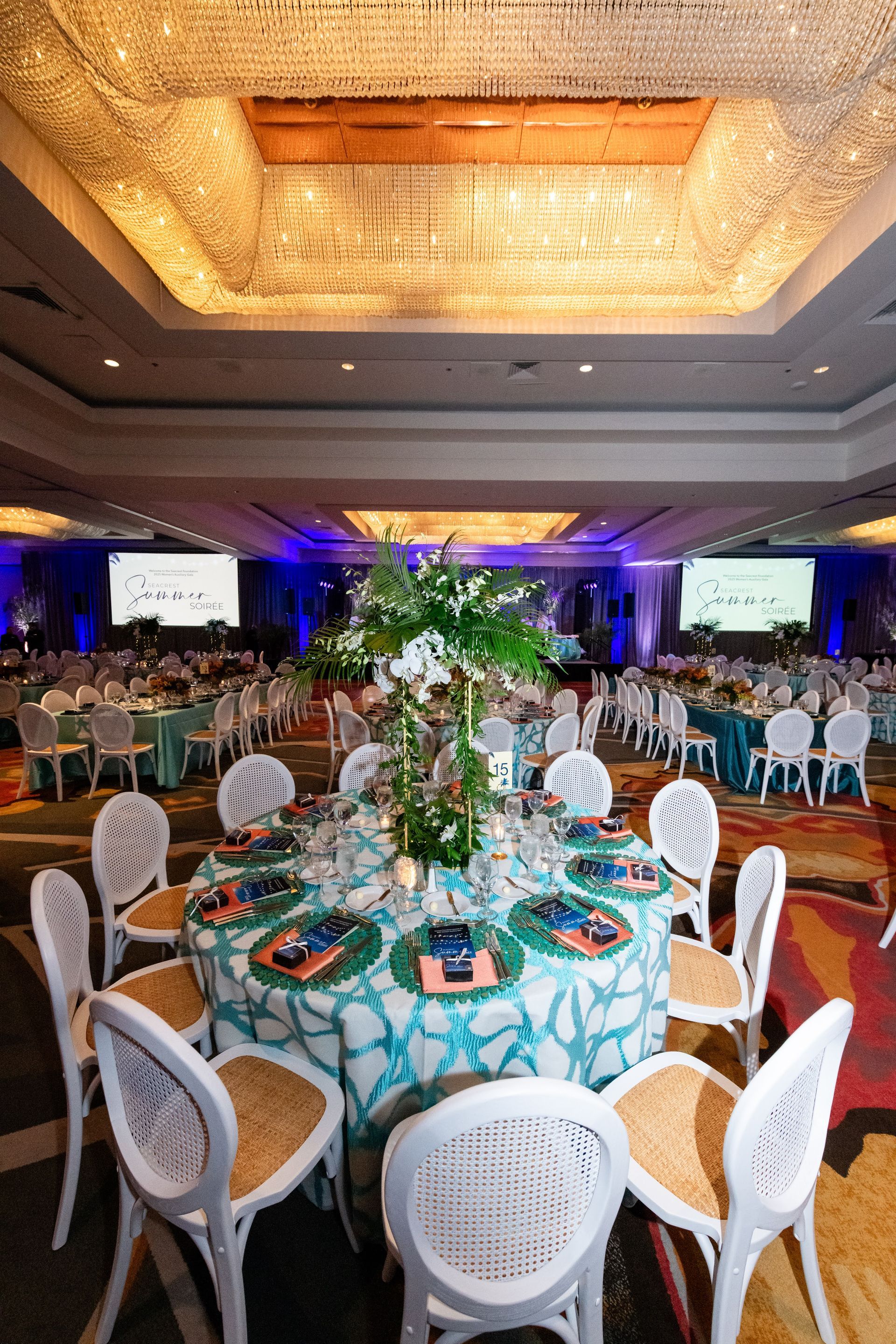 Round table set for a banquet, turquoise tablecloth, white chairs, floral centerpiece, room with blue lighting.