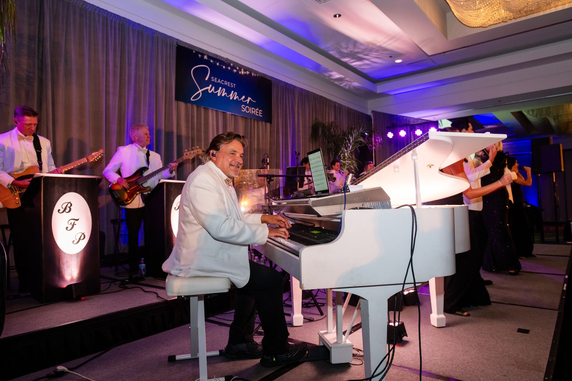 A band performs on stage. A man plays a white piano. Musicians play guitars and sing under blue stage lights.