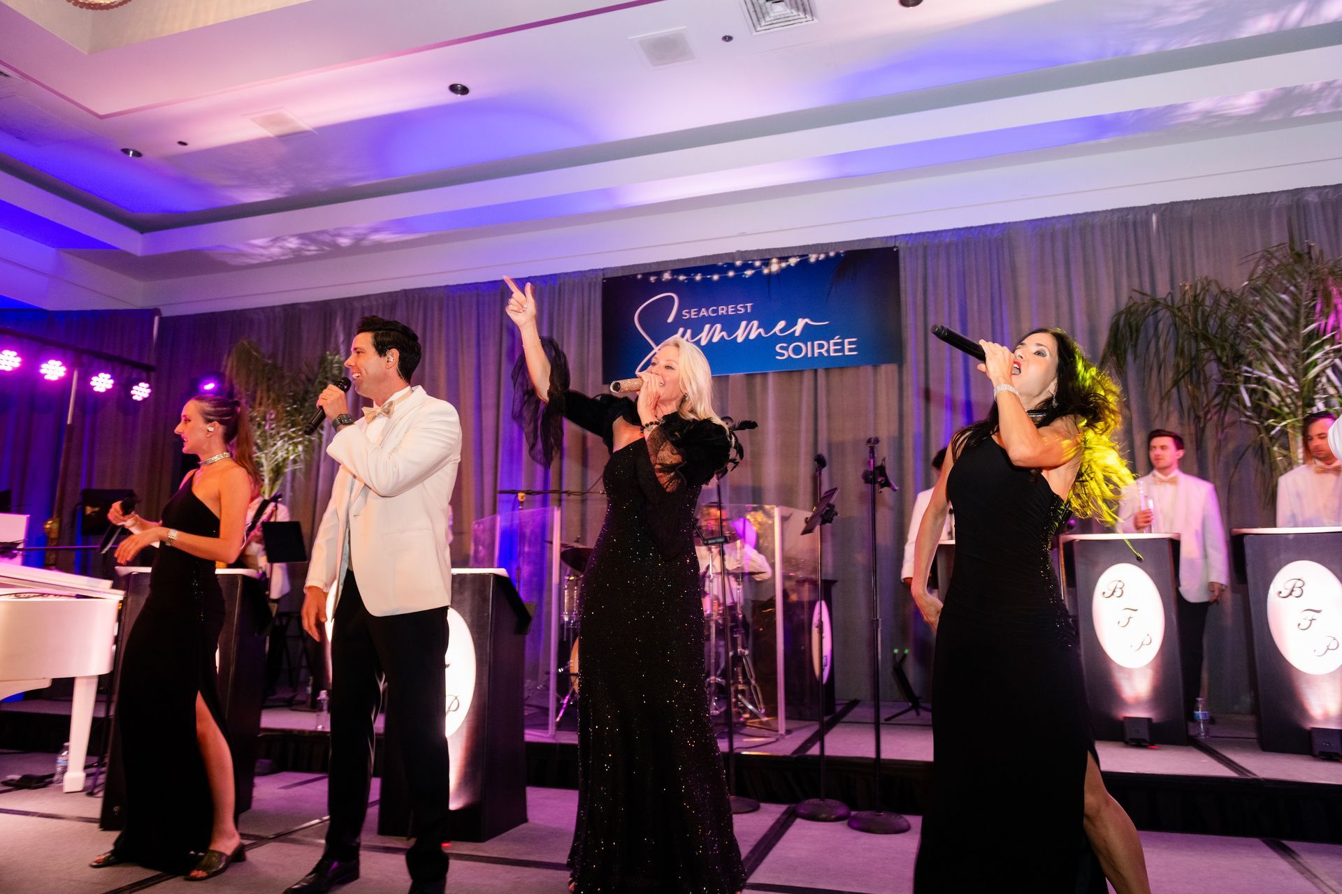 Band performing on stage: vocalists, drums, keyboard, in formal attire, at an event with logo backdrop.