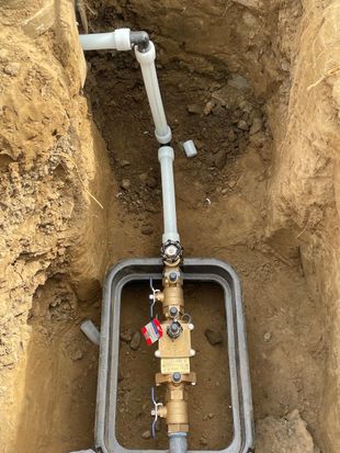 Utility trench with exposed white PVC water pipe and brass shutoff valve in a dirt excavation.