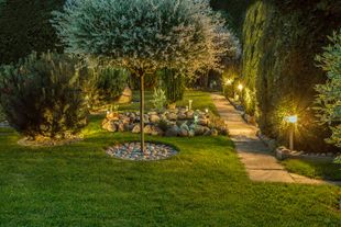 Lit stone garden path leading to a warmly lit patio with sofa seating at dusk.