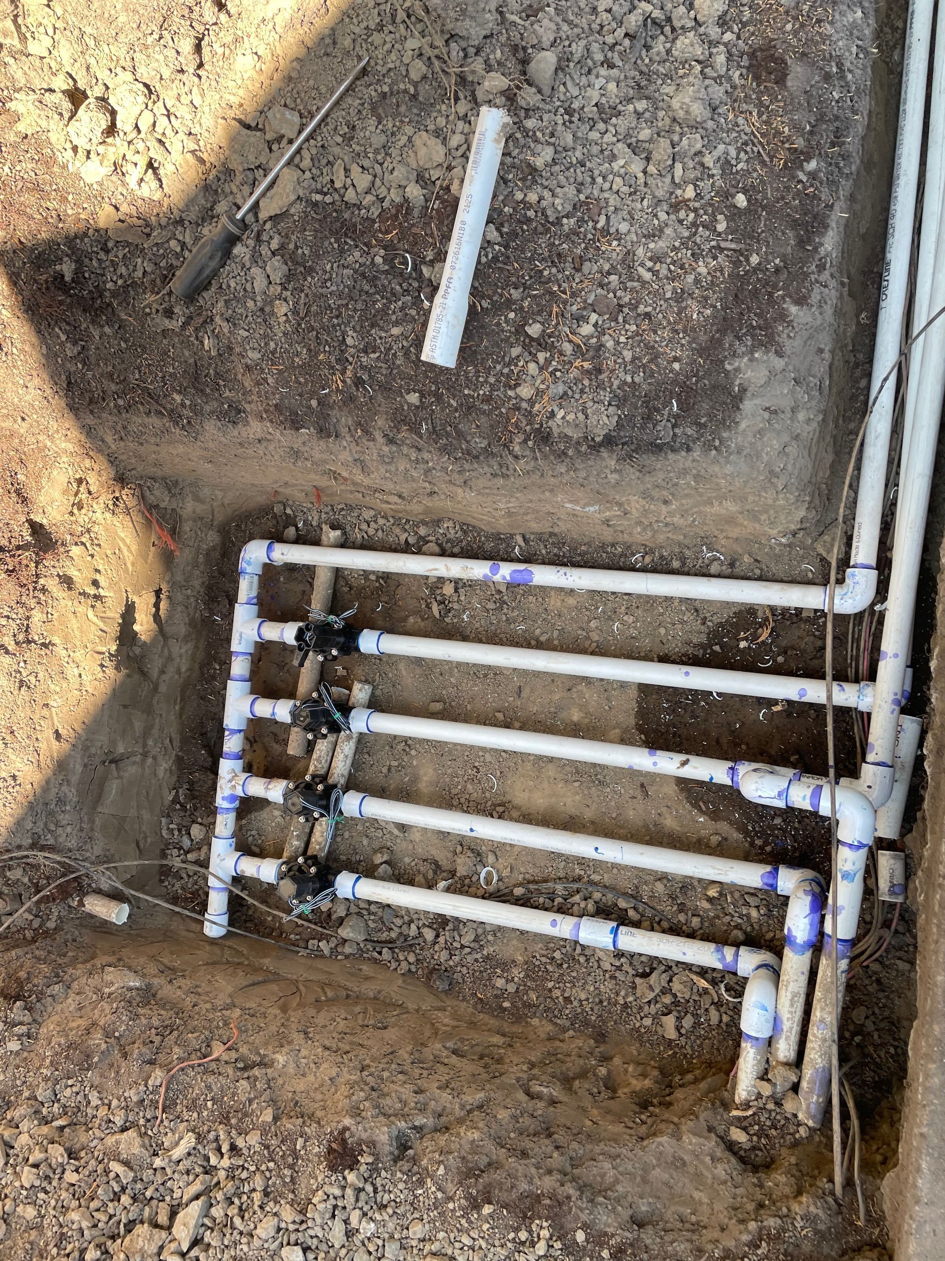 Trenched white PVC pipes and valves laid in dirt, likely an irrigation or plumbing setup.