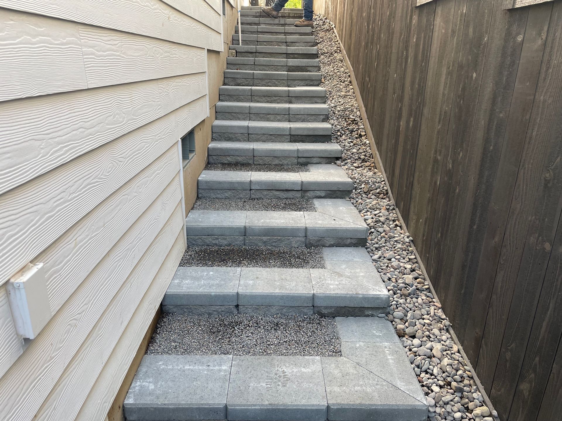 Narrow outdoor concrete steps between a house and a wooden fence, with gravel along the side.