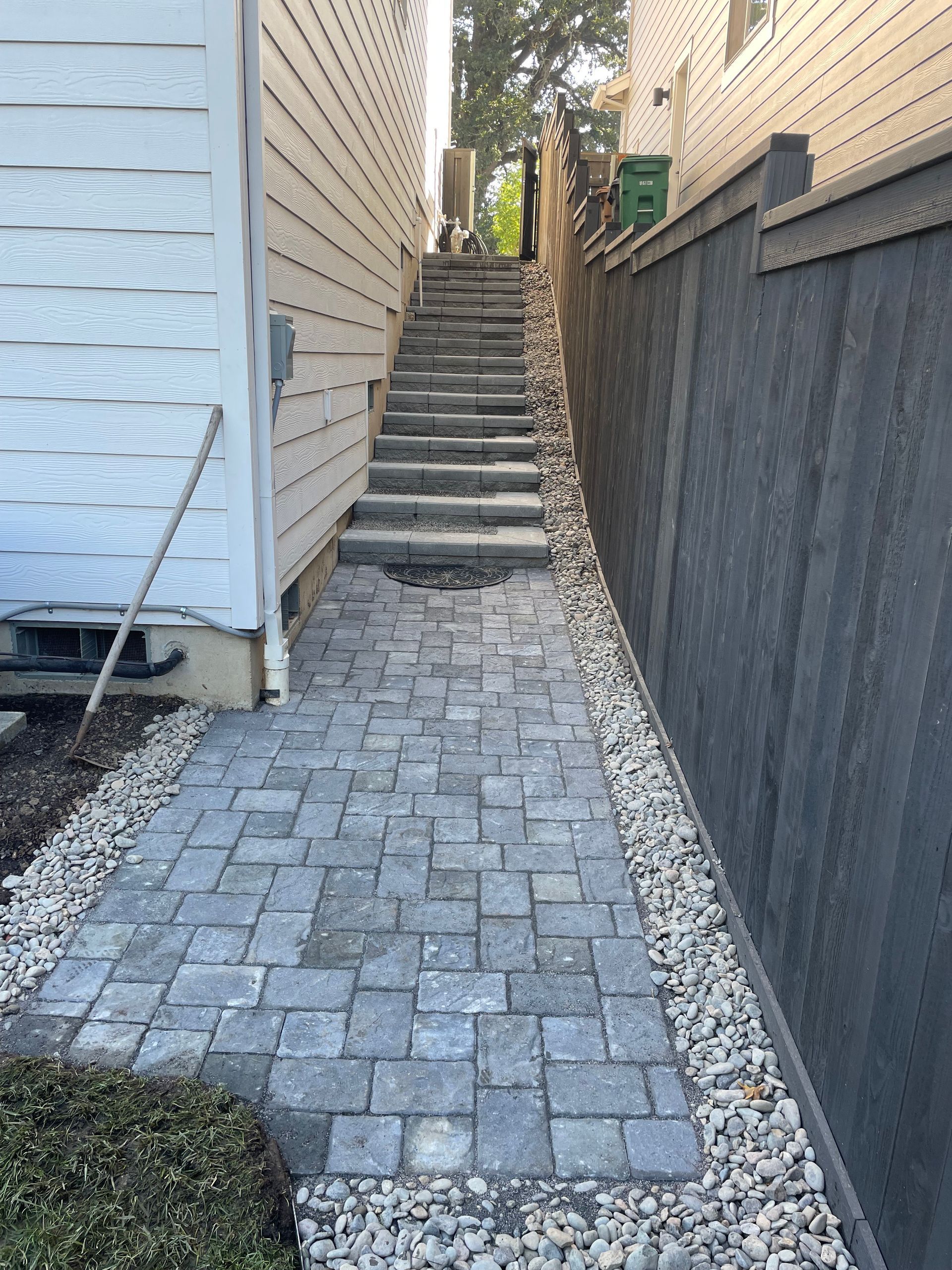 Narrow paved walkway leading up to outdoor concrete steps between two houses, with stone borders and fences.