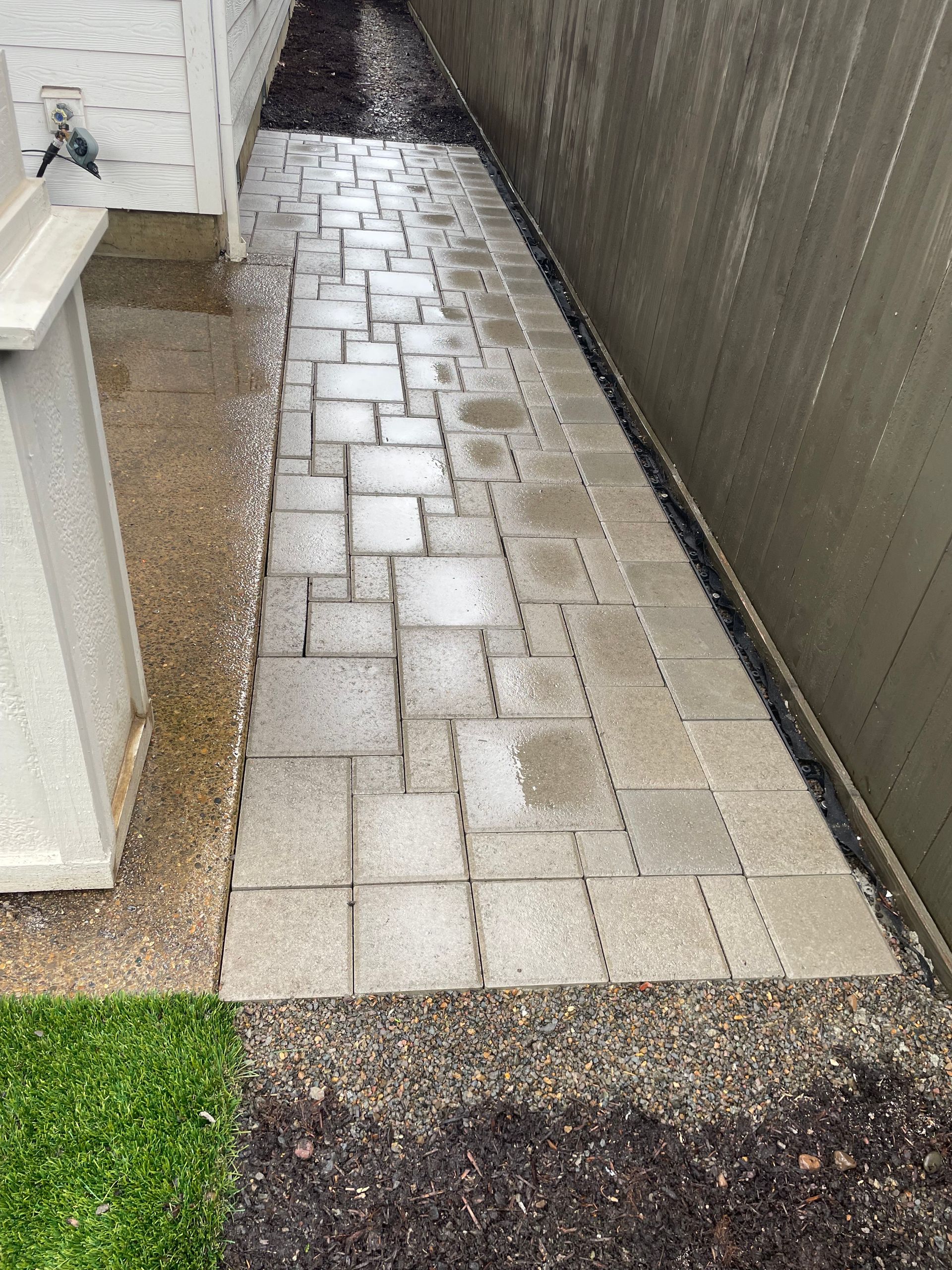 Narrow wet brick walkway between a white house wall and a dark fence, bordered by gravel and grass.