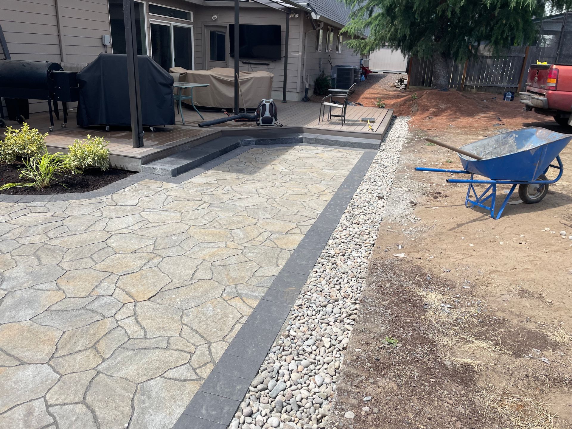 Stone-paved backyard path with a dark border beside a gravel driveway, wheelbarrow, and red truck in a yard.