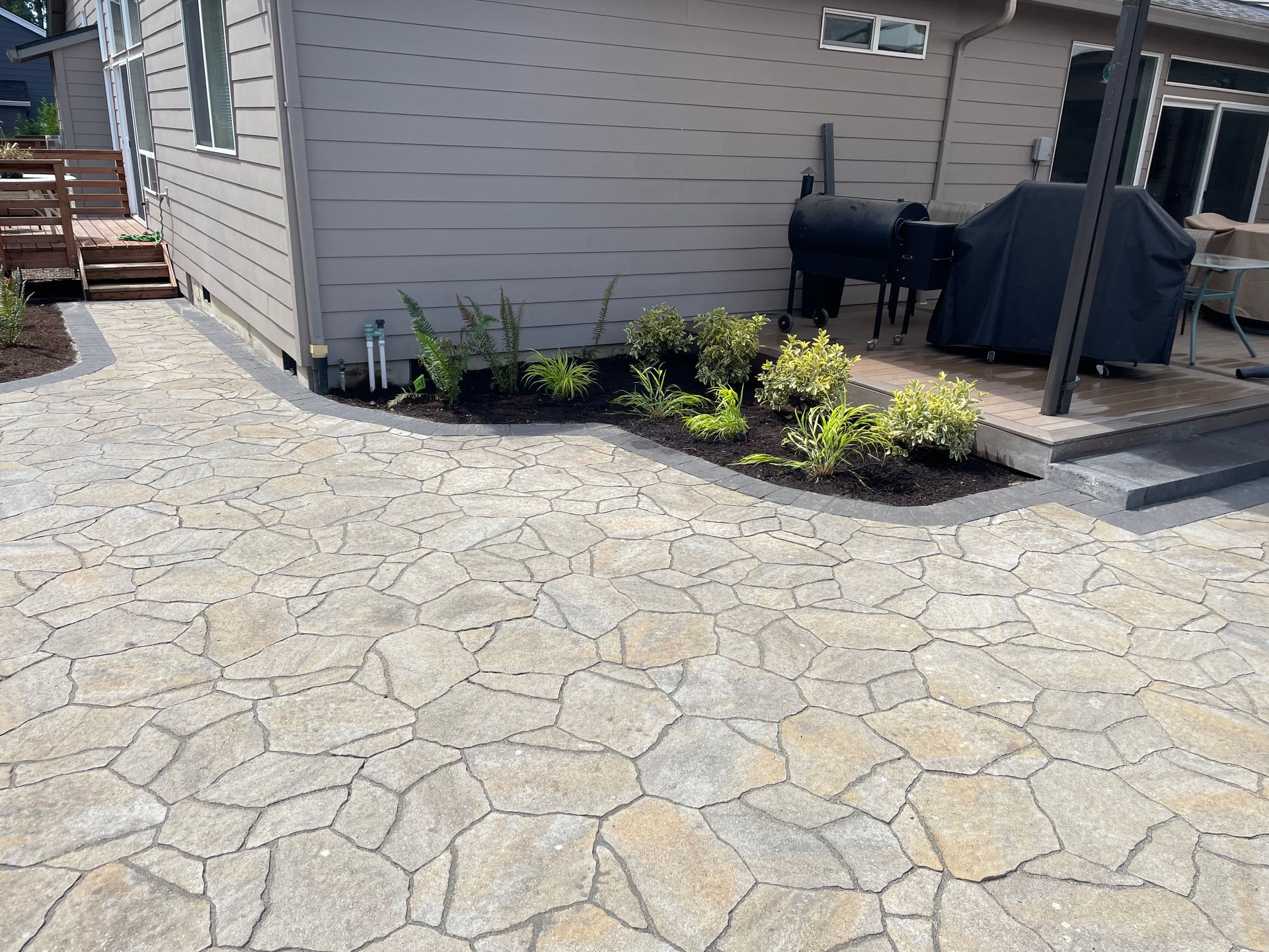 Stone-paver patio beside a house with raised garden beds and a black grill near the steps