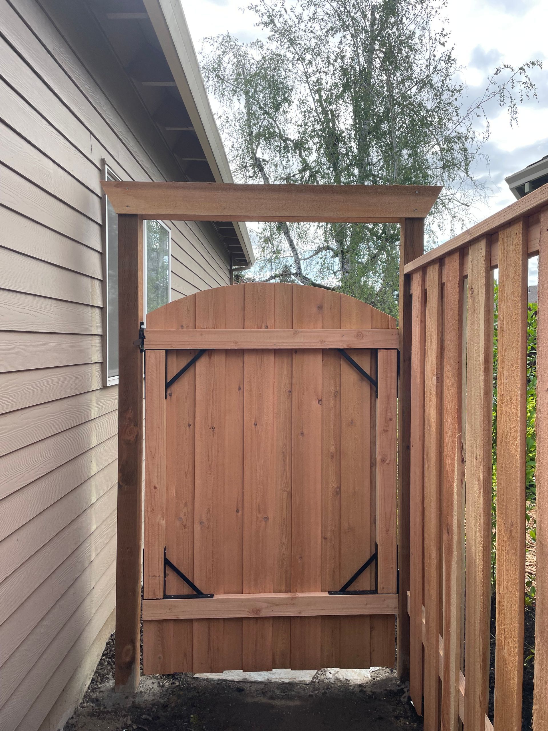 Wooden gate between a house wall and fence, with a small overhead frame and trees behind.