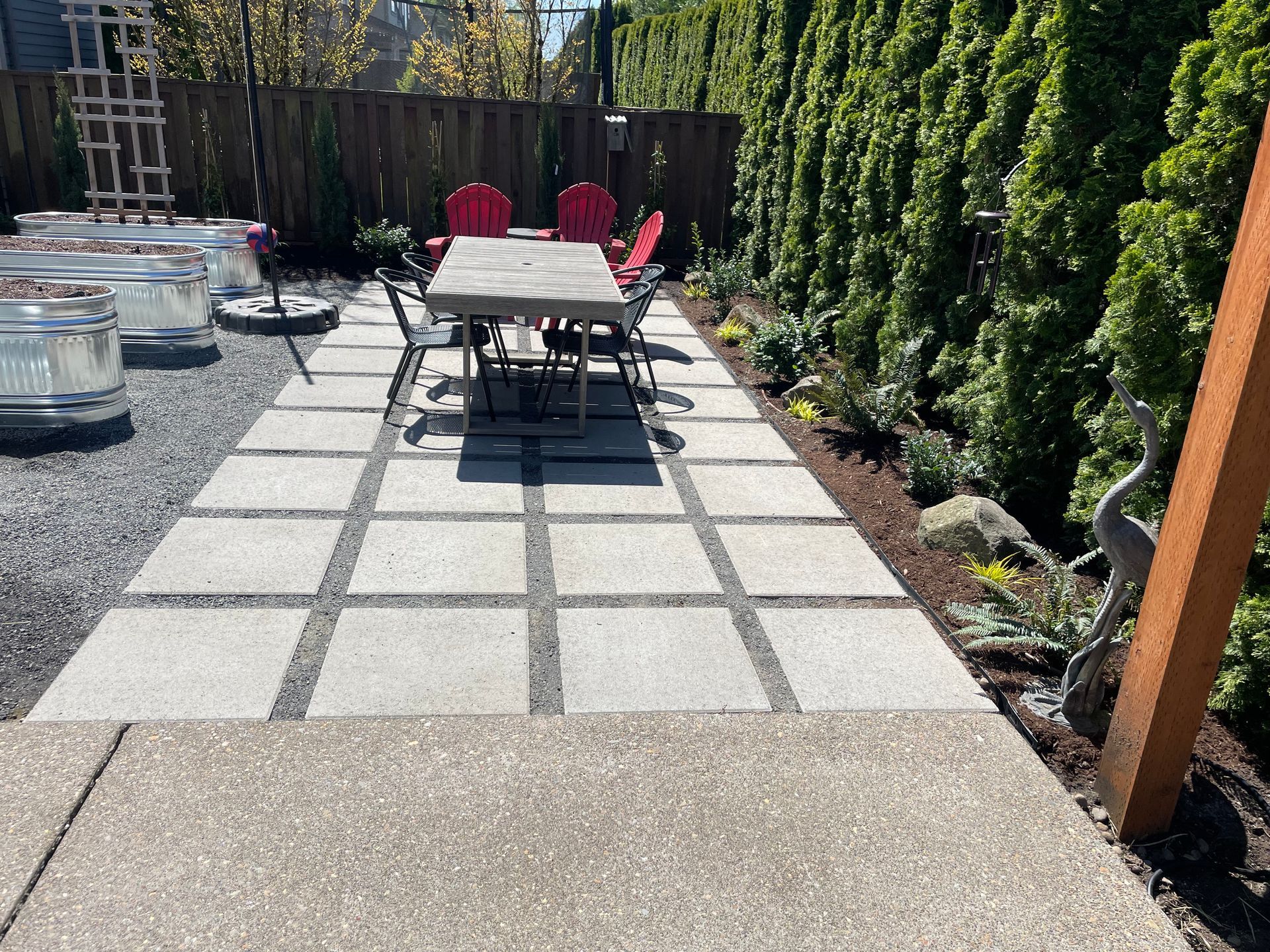 Patio with square pavers, outdoor dining table and chairs, red loungers, and a gravel path beside greenery