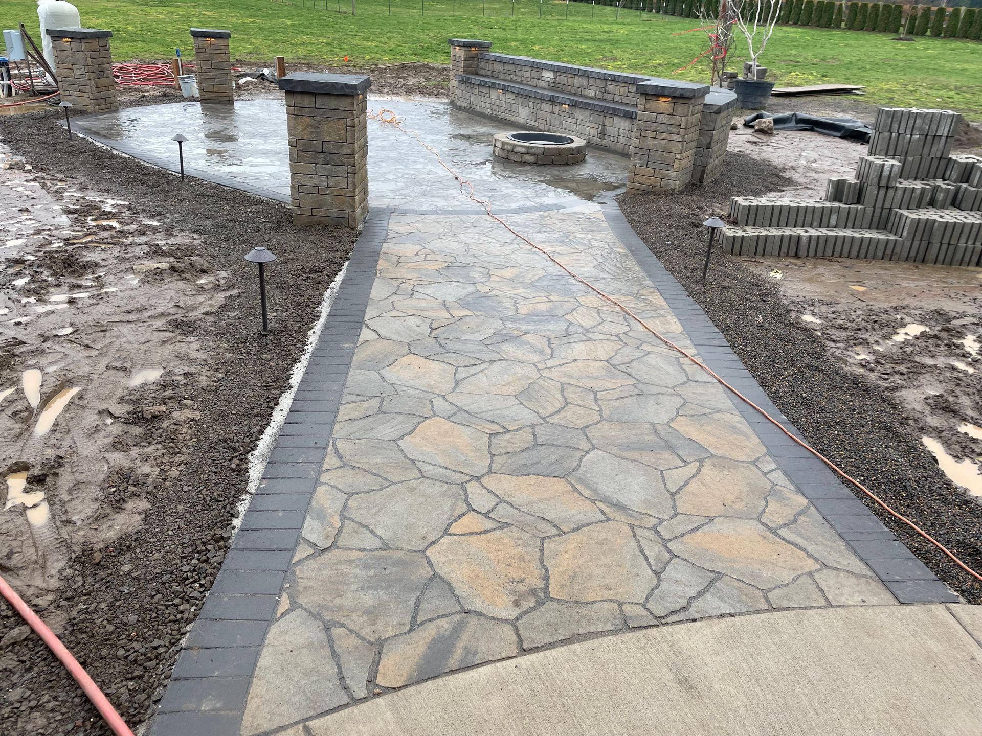 Stone walkway under construction beside a muddy yard and low retaining wall in a backyard.