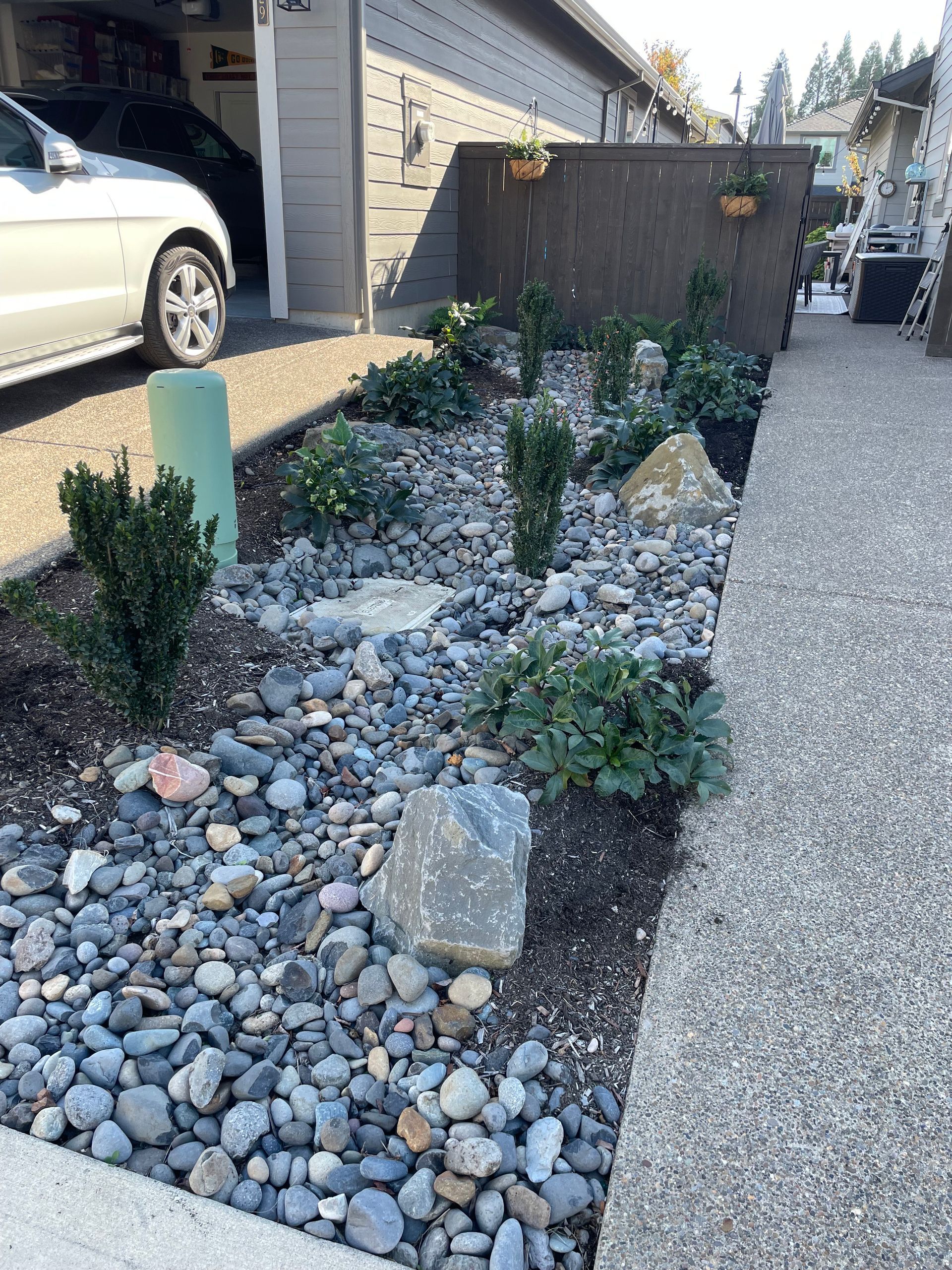 Gravel and rock landscaped garden bed beside a driveway and house wall, with small shrubs and a concrete retaining wall.