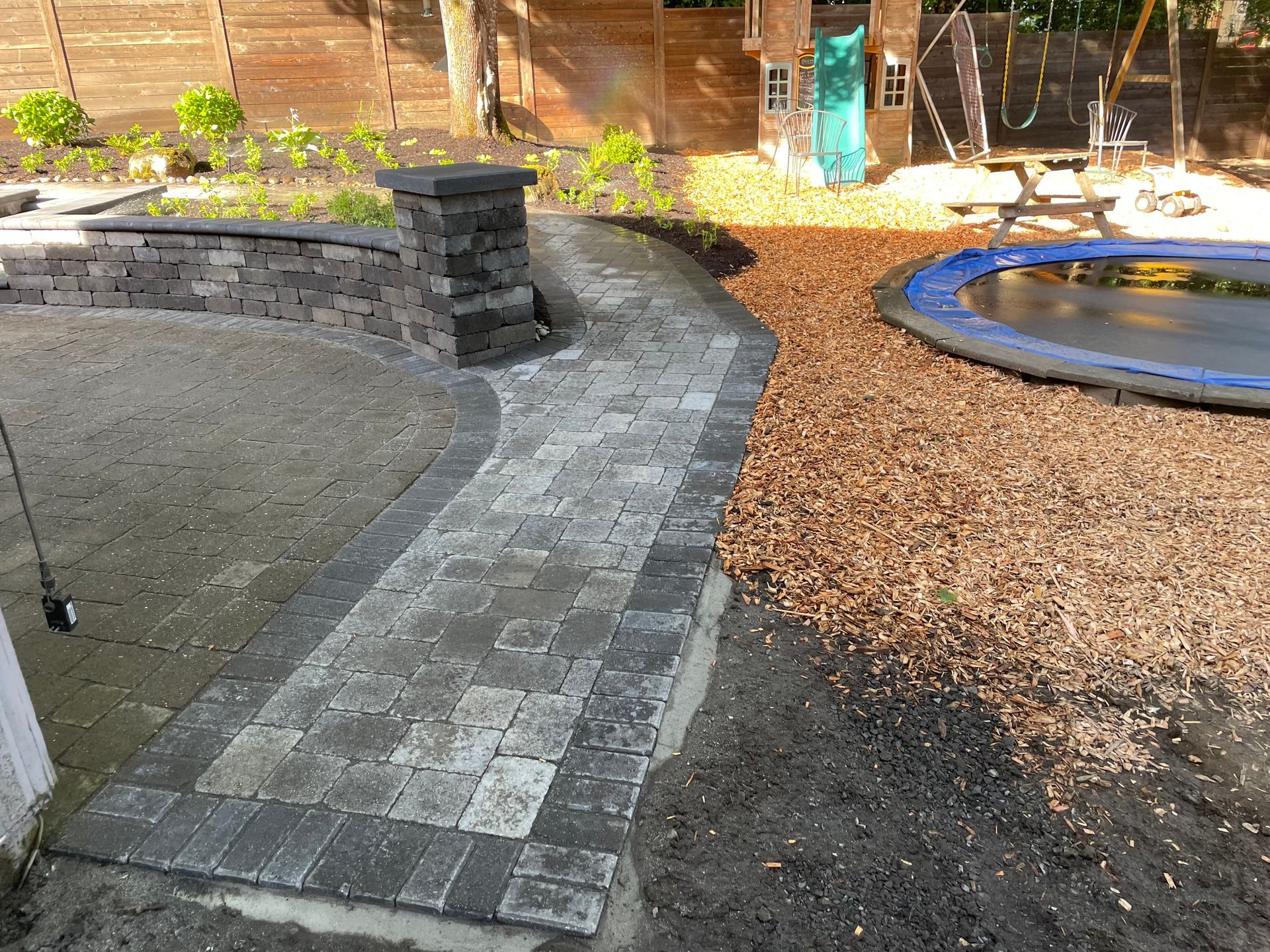 Backyard stone path beside a small pond and gravel area, with a brick retaining wall and fence