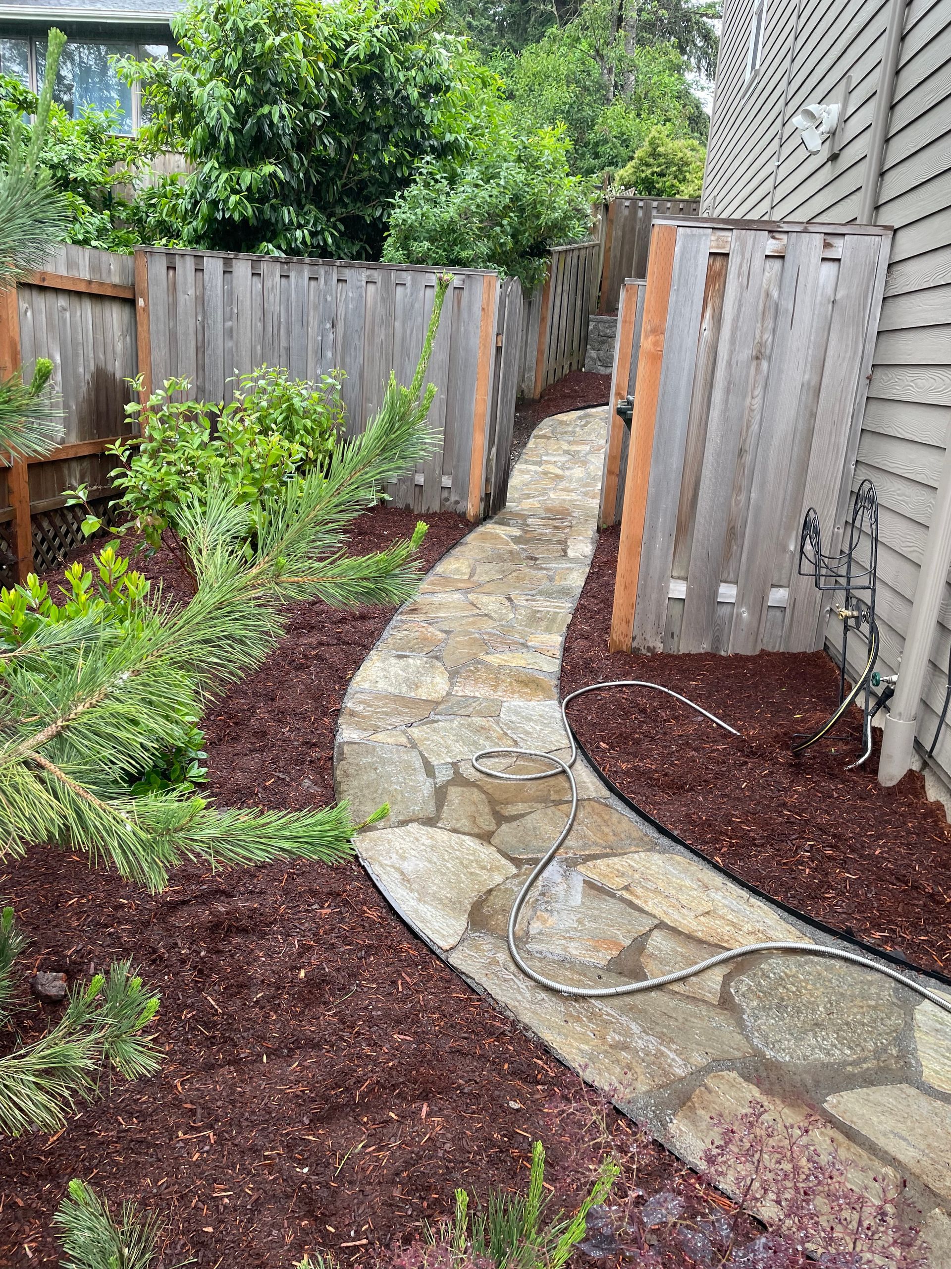 Garden path with stone pavers, mulch beds, and wooden fences beside a house wall