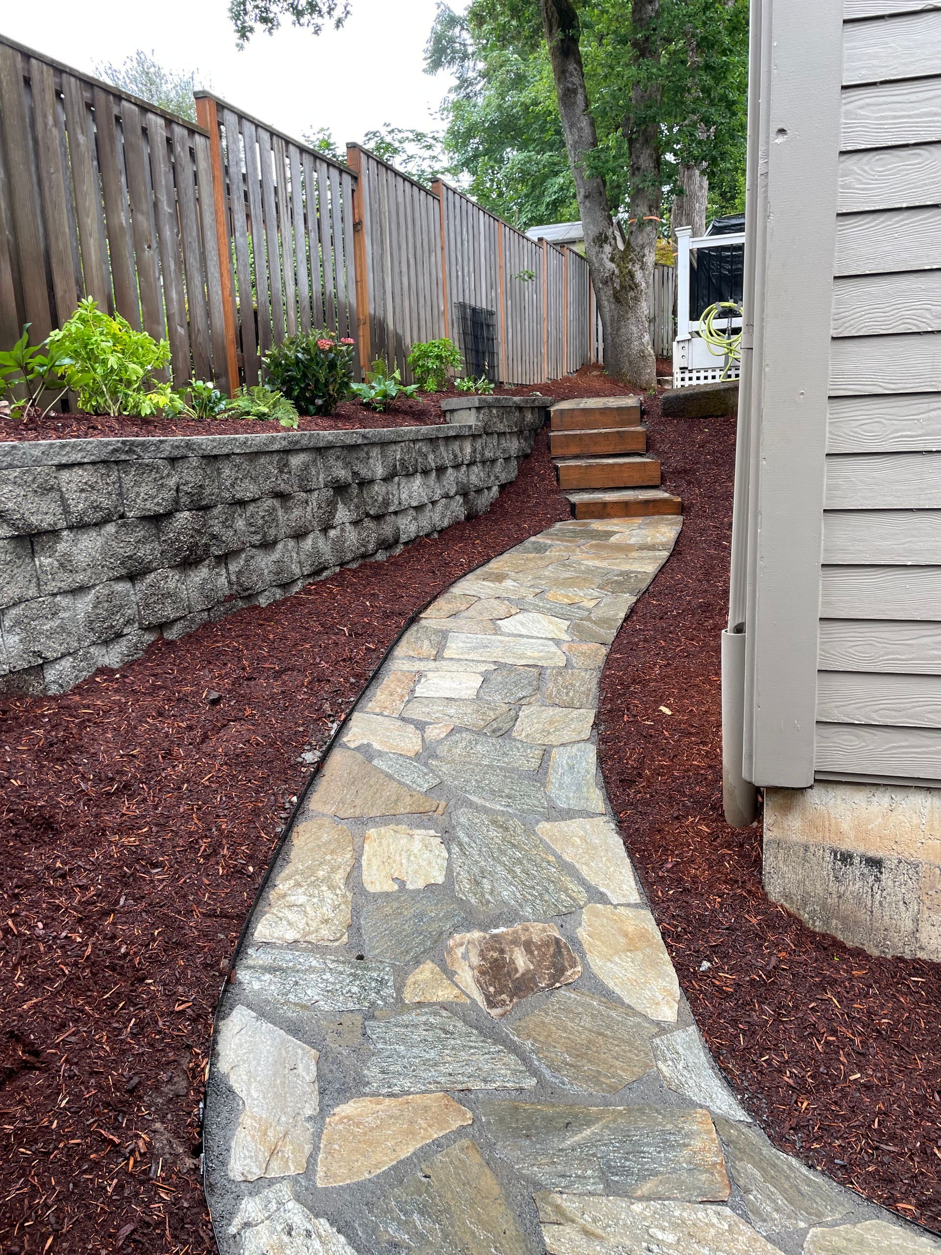 Curved stone pathway beside a raised rock garden and wooden fence in a narrow side yard