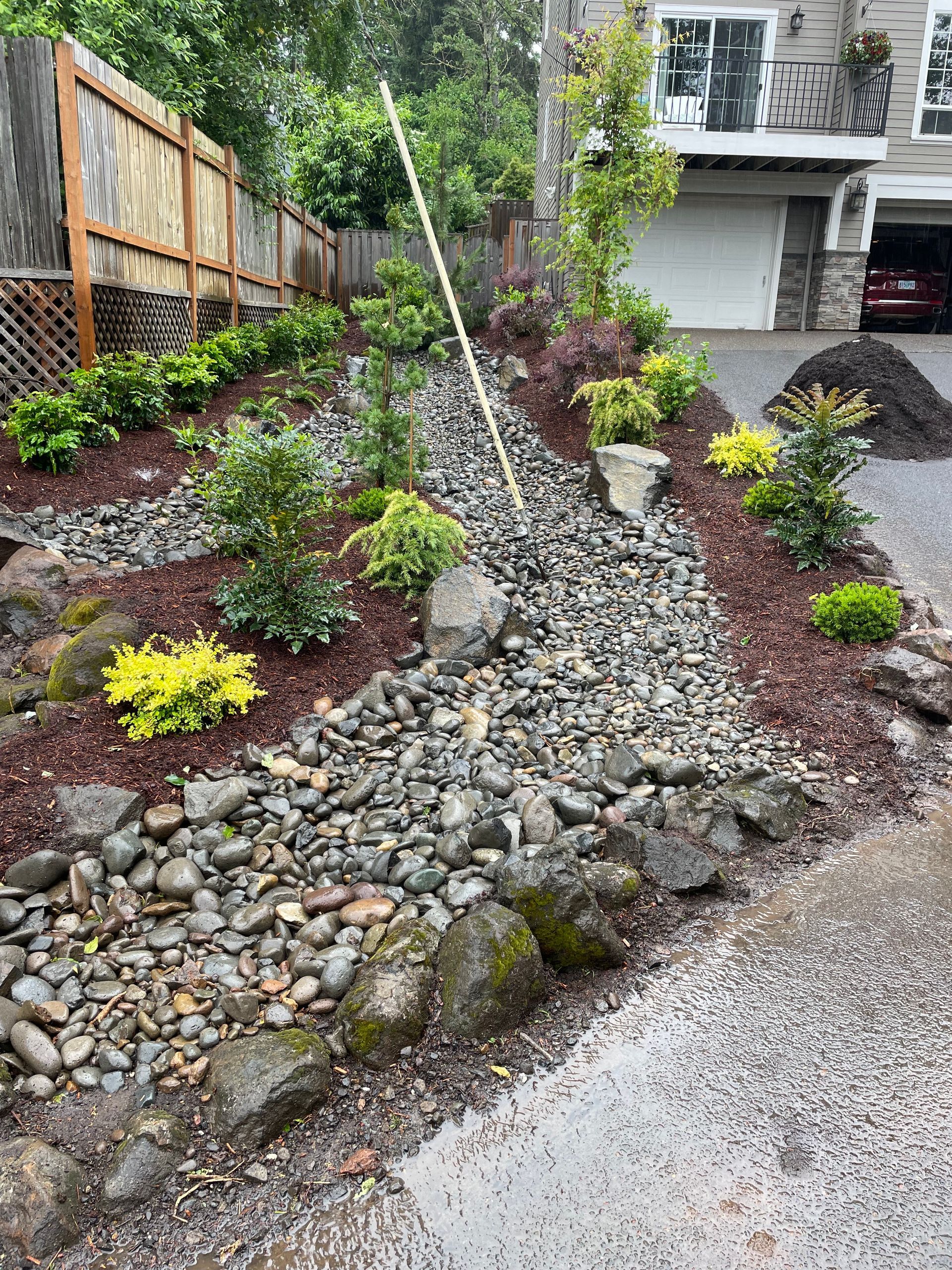Landscaped front yard with rock drainage bed, small shrubs, and a house entrance in the background