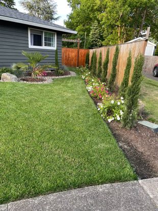 Front yard with lush lawn, gray house, flower bed, wooden fence, and a paved driveway edge.