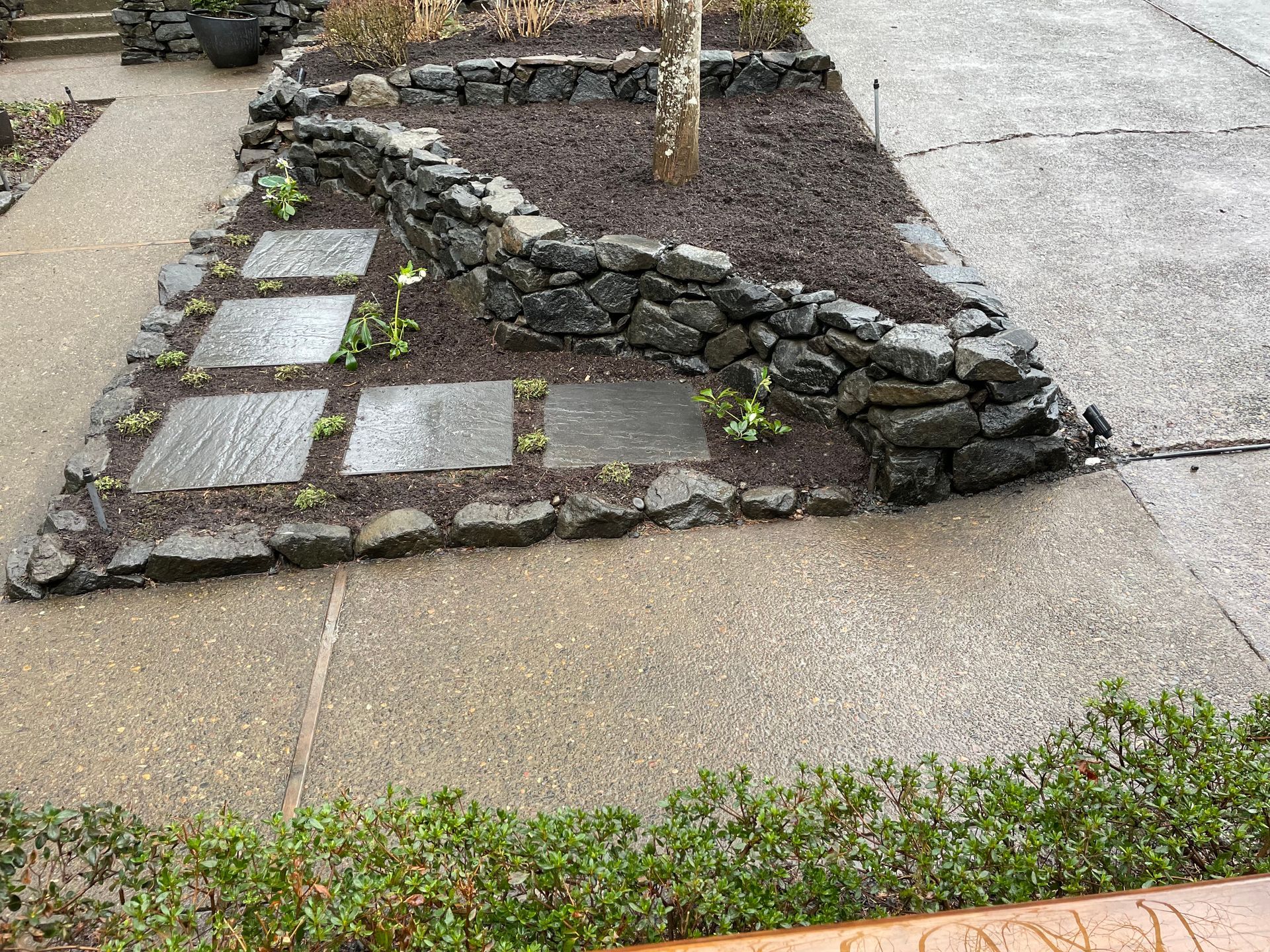 Stone garden border with square stepping stones beside a concrete path and a low wall of rocks