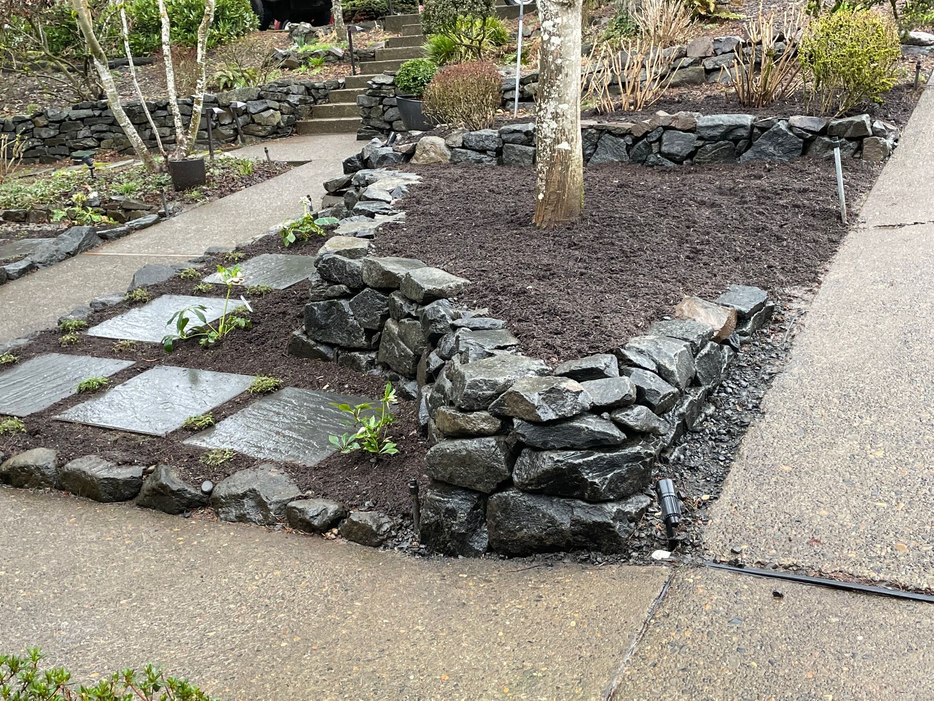 Stone-edged garden bed with stepping stones and dark mulch beside a walkway