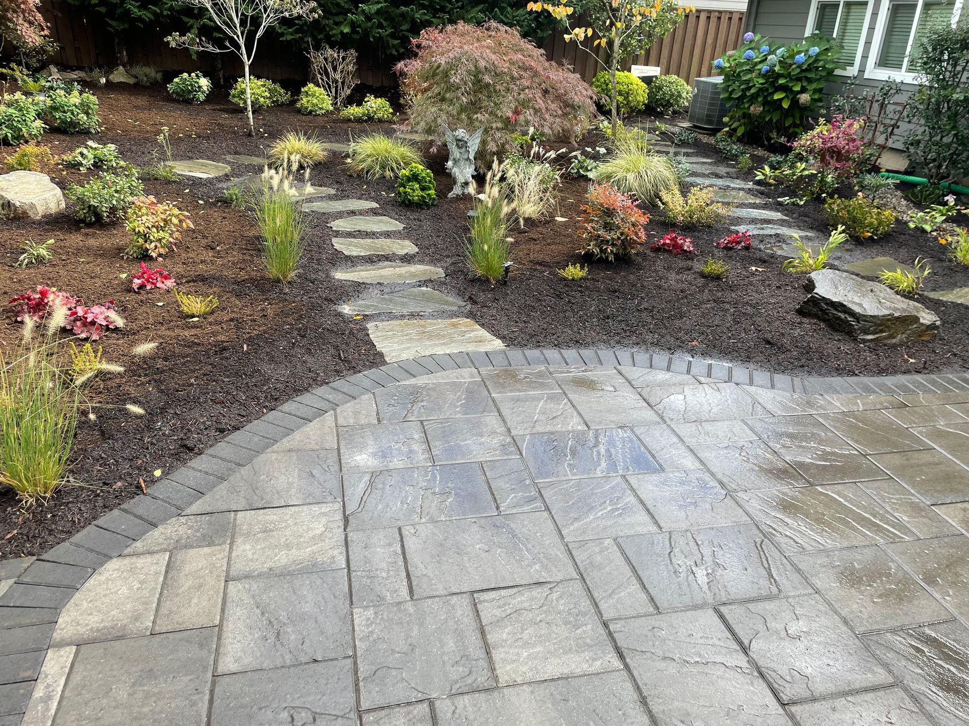 Curved stone patio leading to a landscaped garden with shrubs, flowers, and stepping-stone path