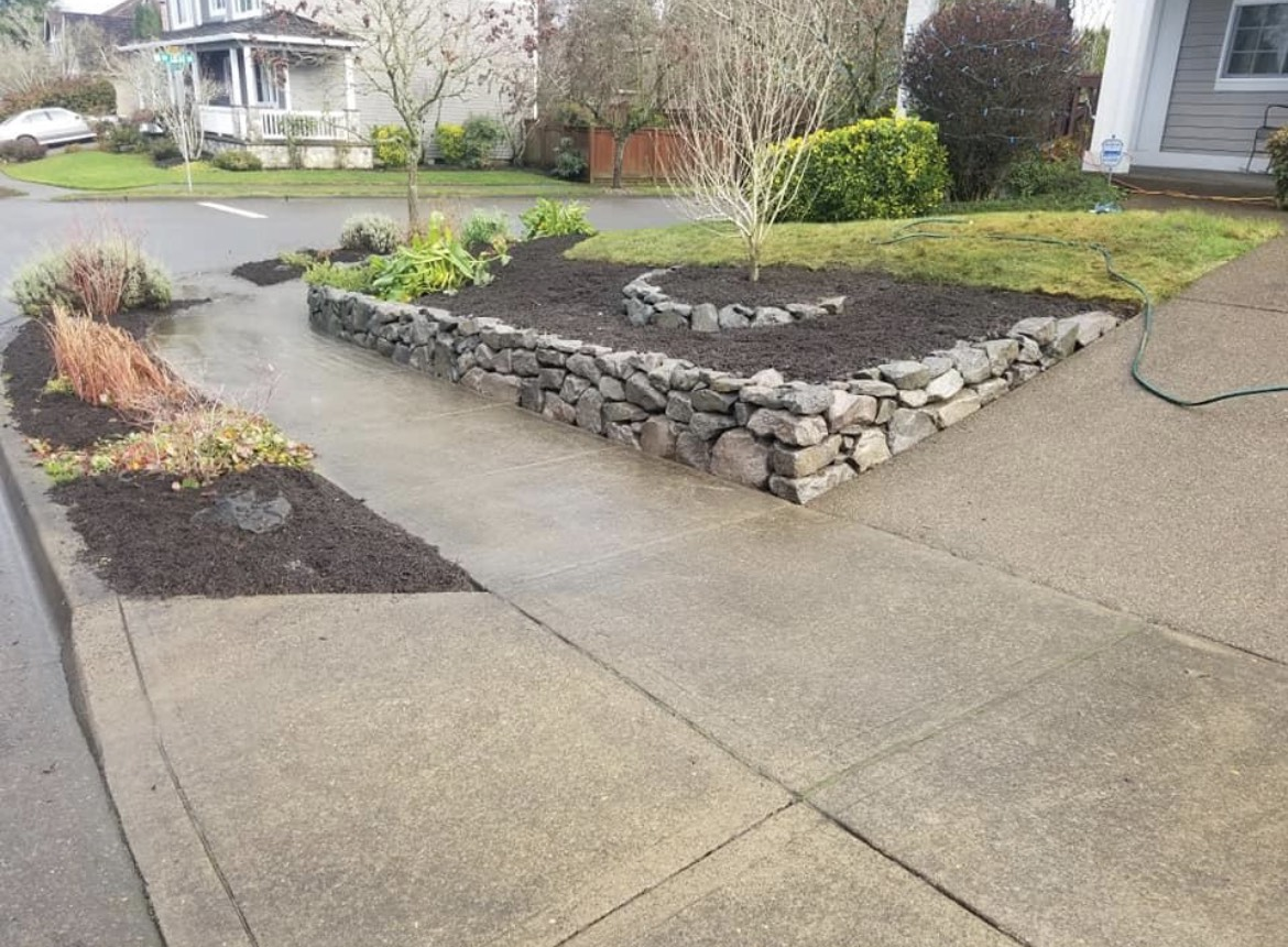 Concrete driveway with stone-edged raised planter beds and shrubs beside a house