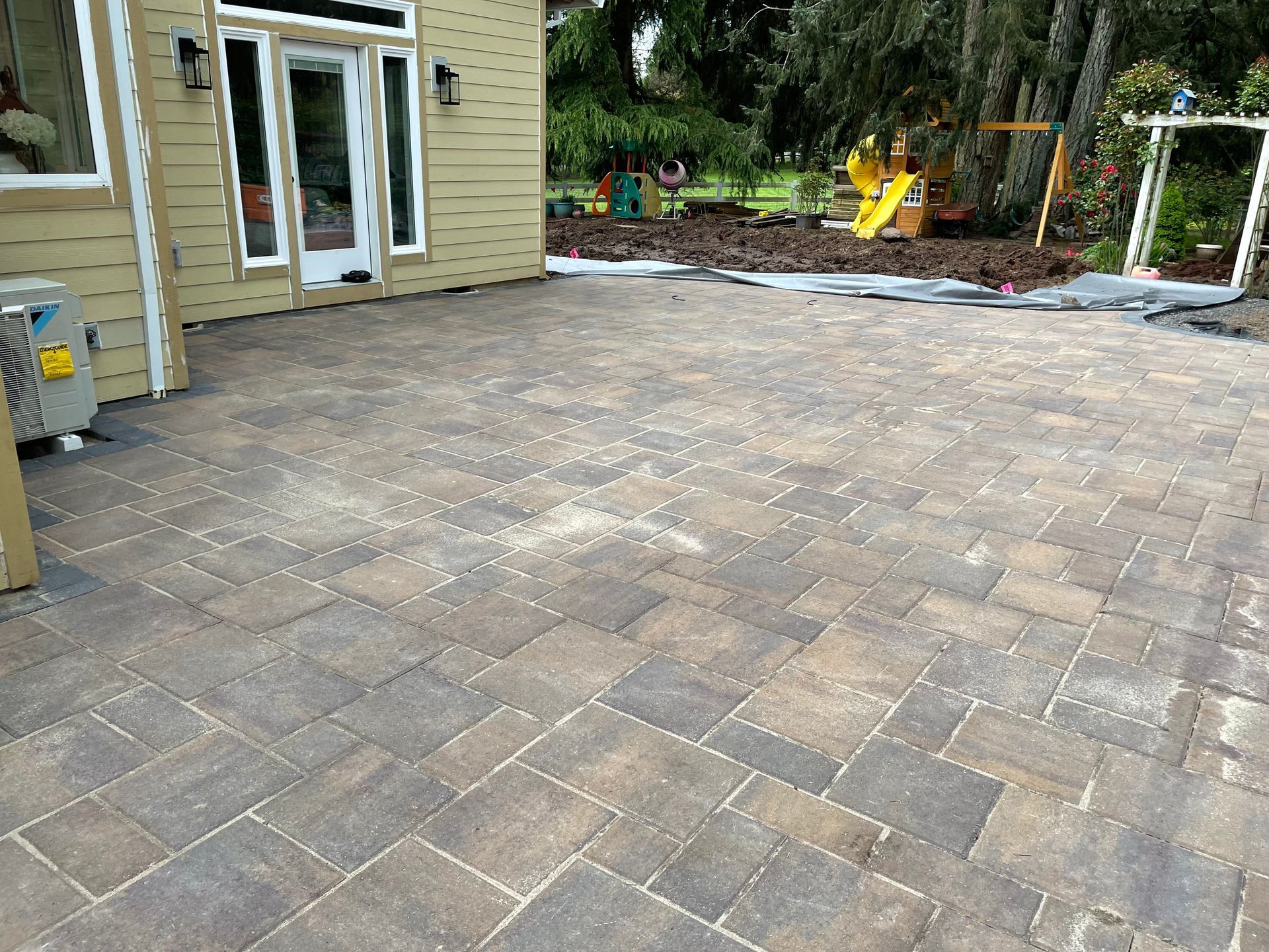 Backyard patio with stone pavers beside a house, leading to a garden area and yellow chair set