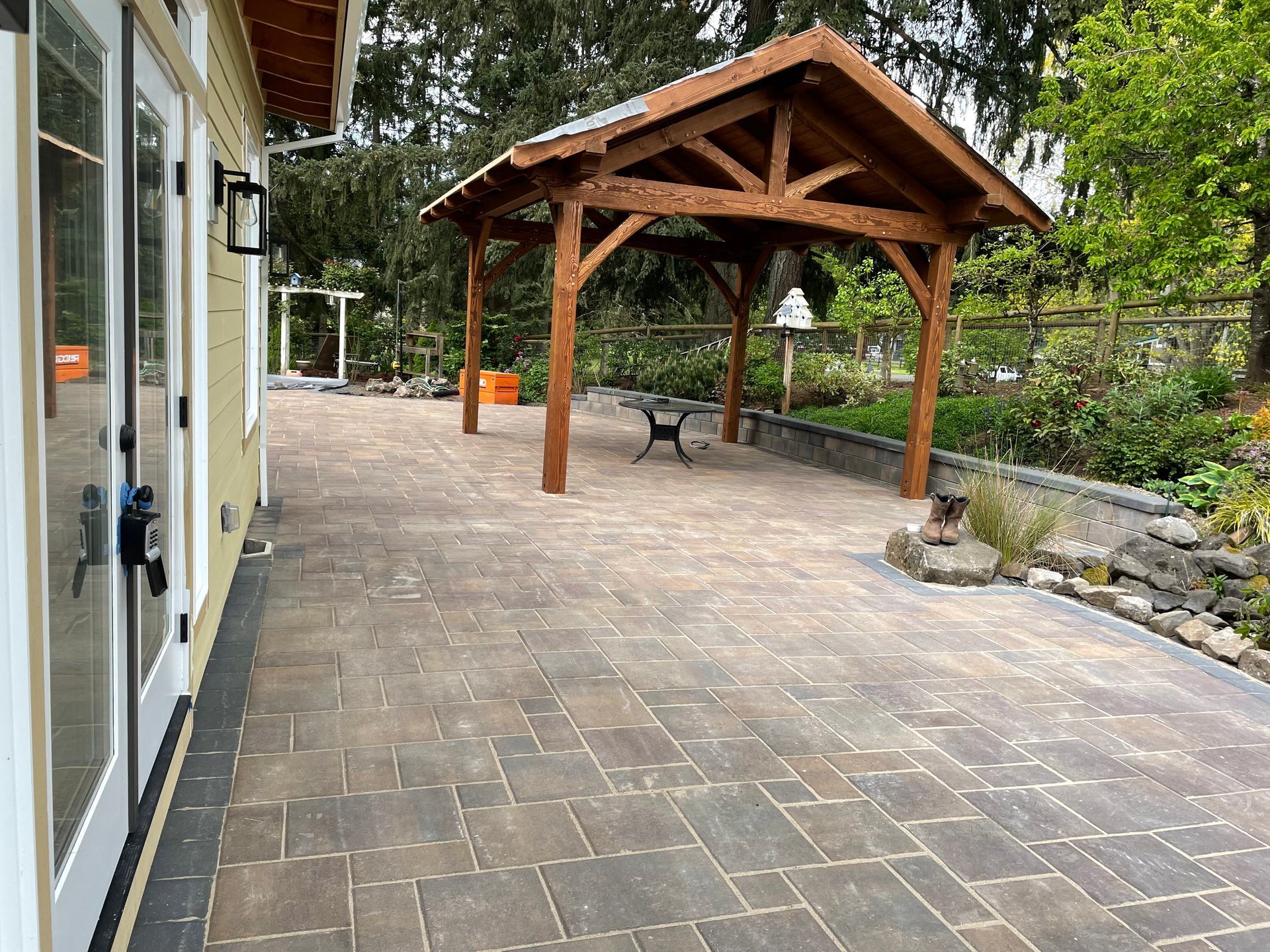 Stone patio with a wooden pavilion and bench beside a building and garden path