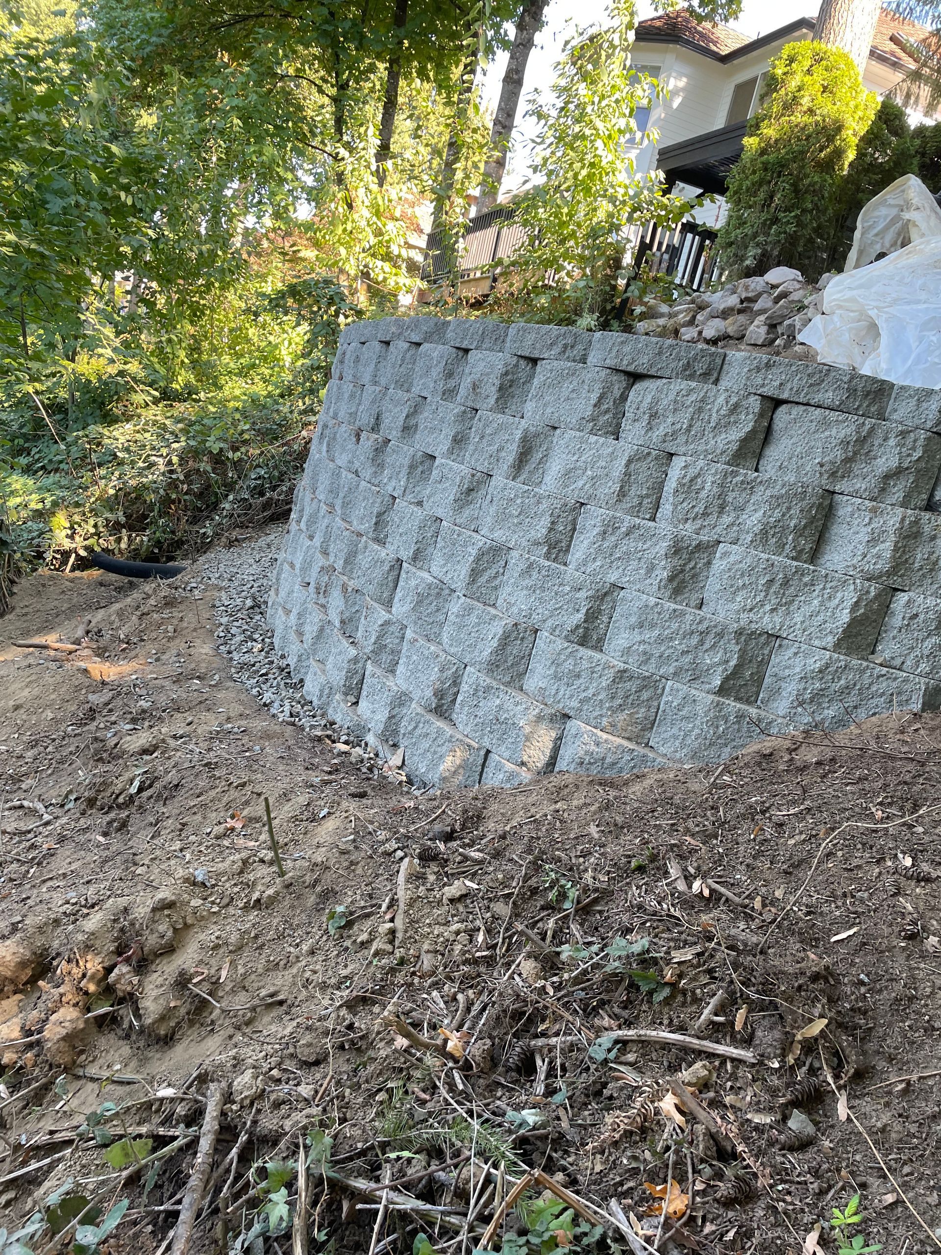 Steep dirt slope beside a gray retaining wall, with trees and a house partially visible above.