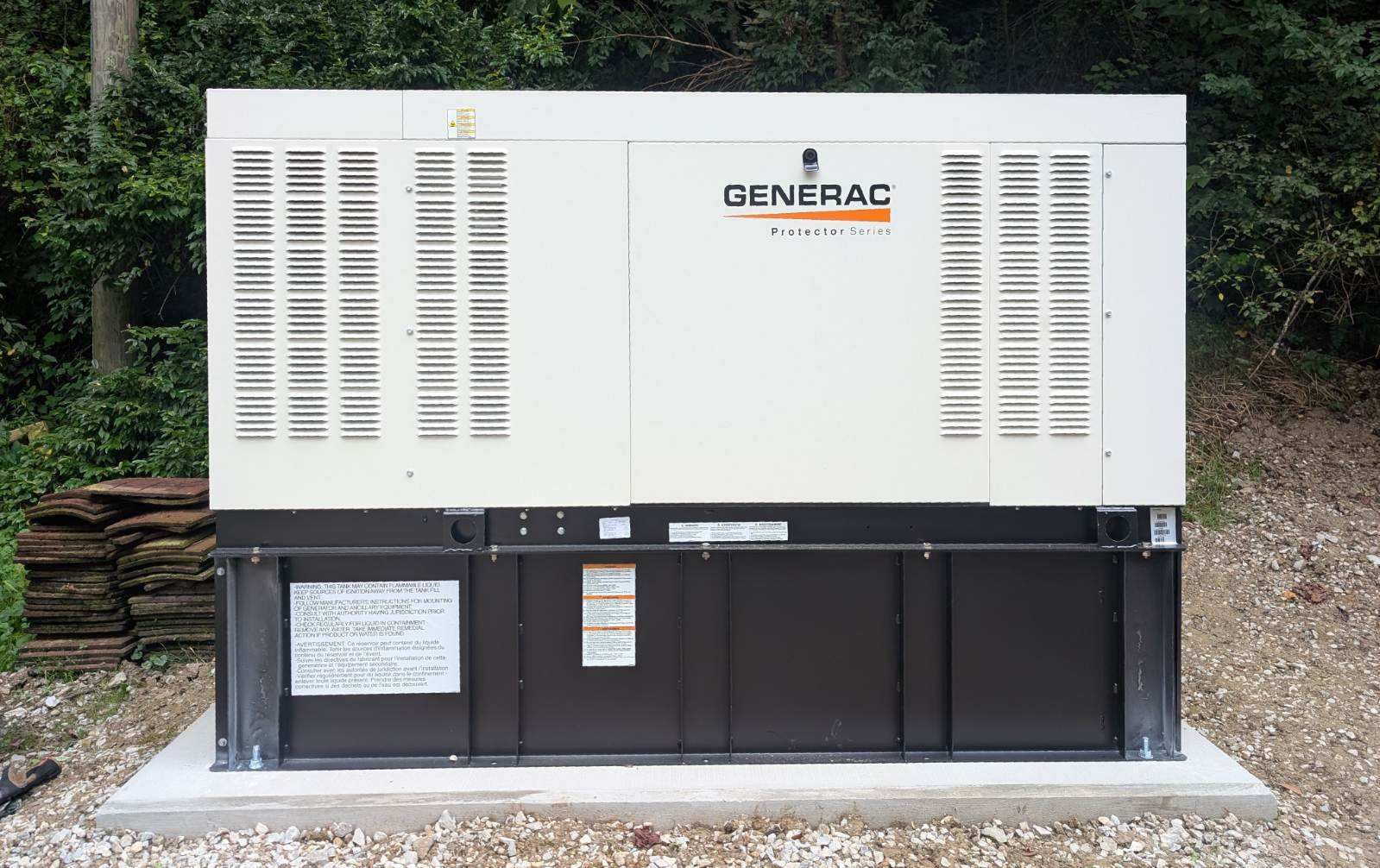 A Generac industrial standby generator sits on a concrete pad outdoors, surrounded by a gravel area and wooded landscape.