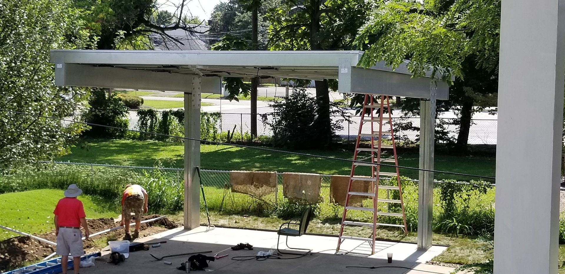 Men working on a metal carport in a yard with a ladder, chairs, and tools.