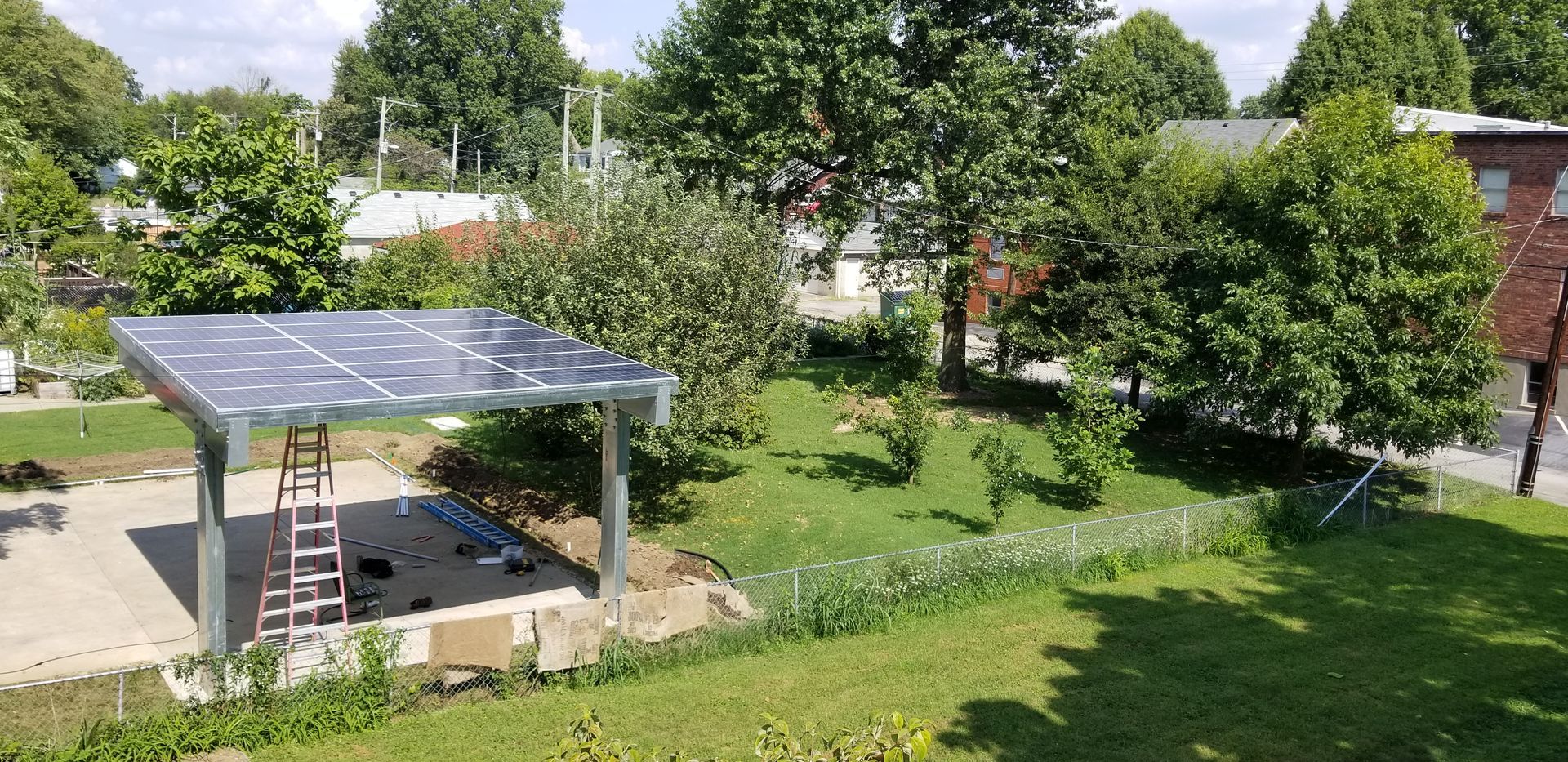Solar panel carport in a yard with trees and grass. A ladder leans against the structure.