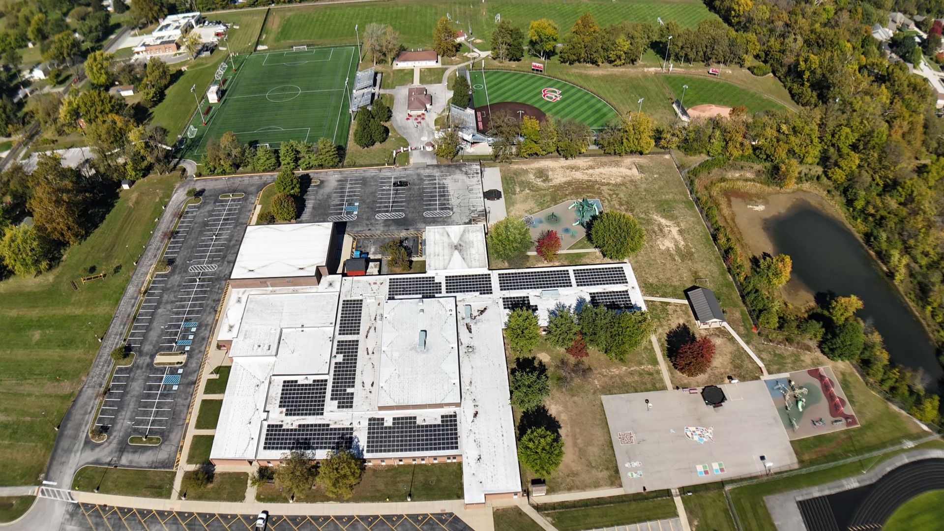 Aerial view of a school campus with buildings, parking lot, sports fields, trees, and a pond.