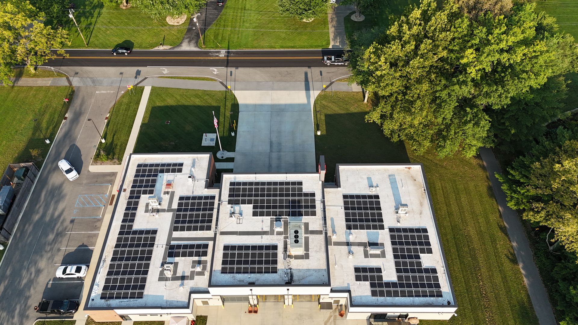 Aerial view of a building with solar panels on the roof and a road alongside. Green trees and grass surround the structure.