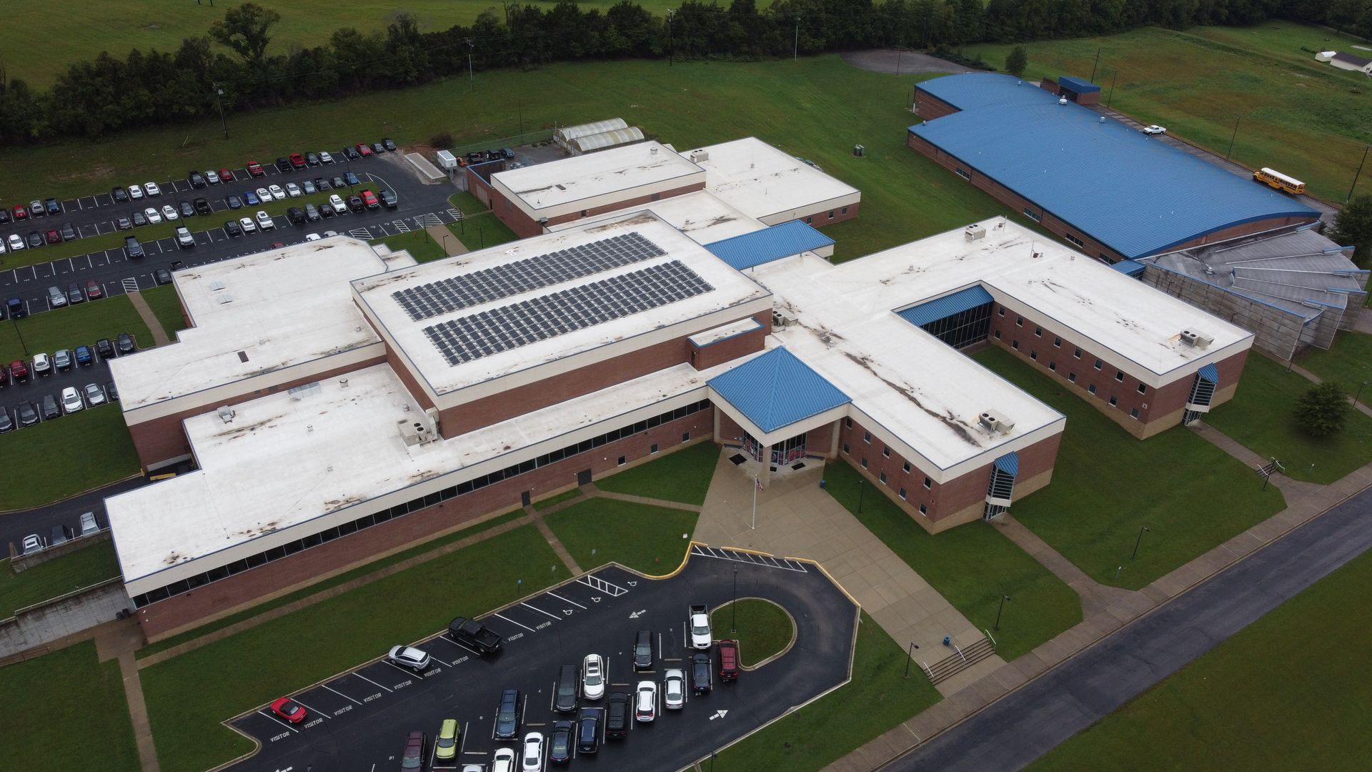 Aerial view of a school building with solar panels on the roof, surrounded by parking lots and a field.