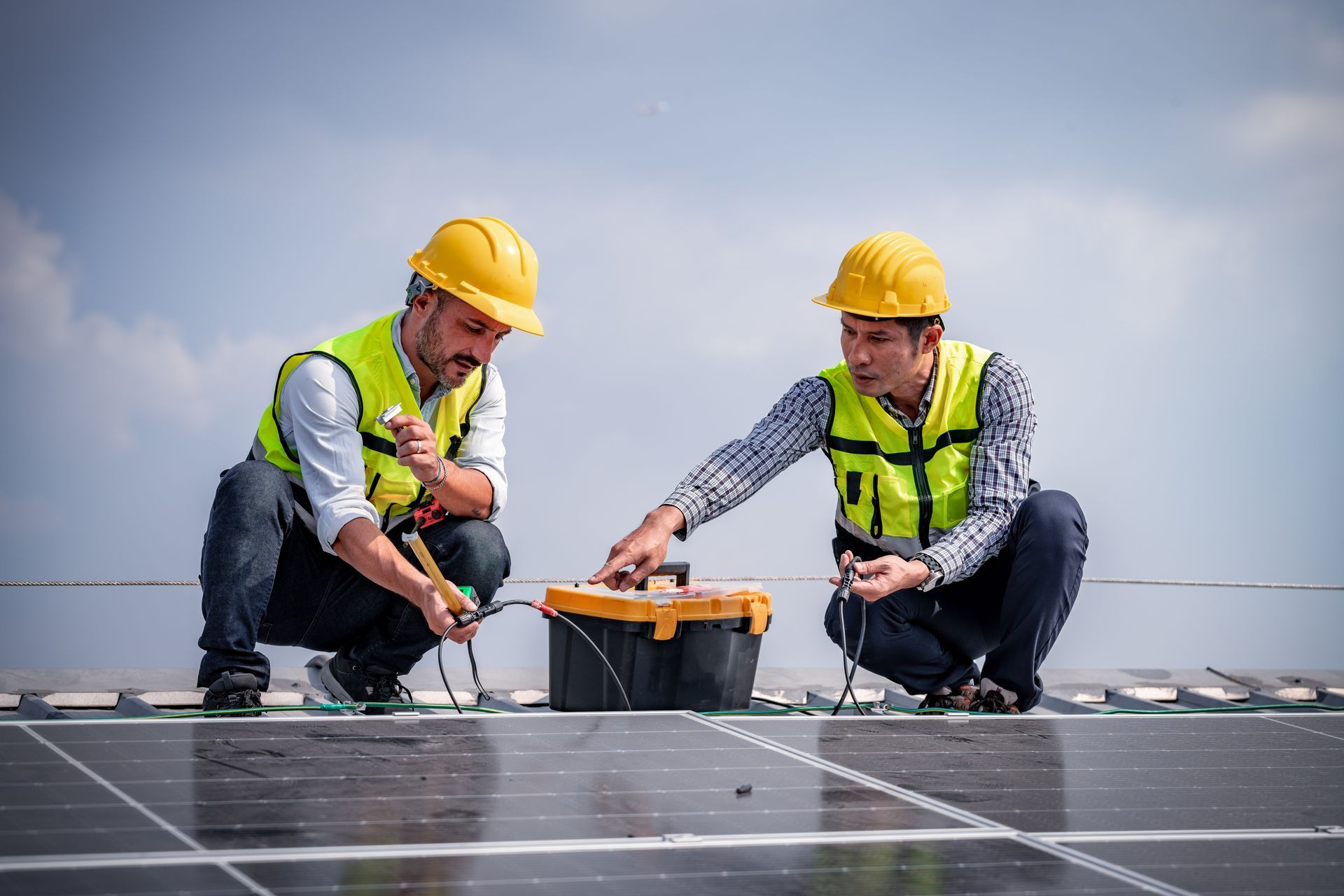 Two workers in hard hats inspect solar panels on a rooftop using a tool box.