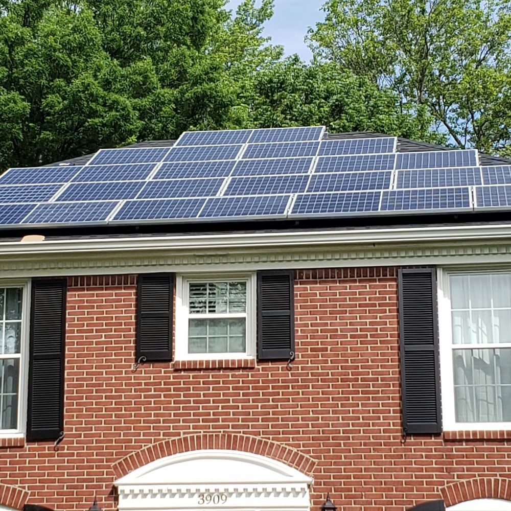 Solar panels on the roof of a brick house with black shutters and windows, surrounded by green trees.