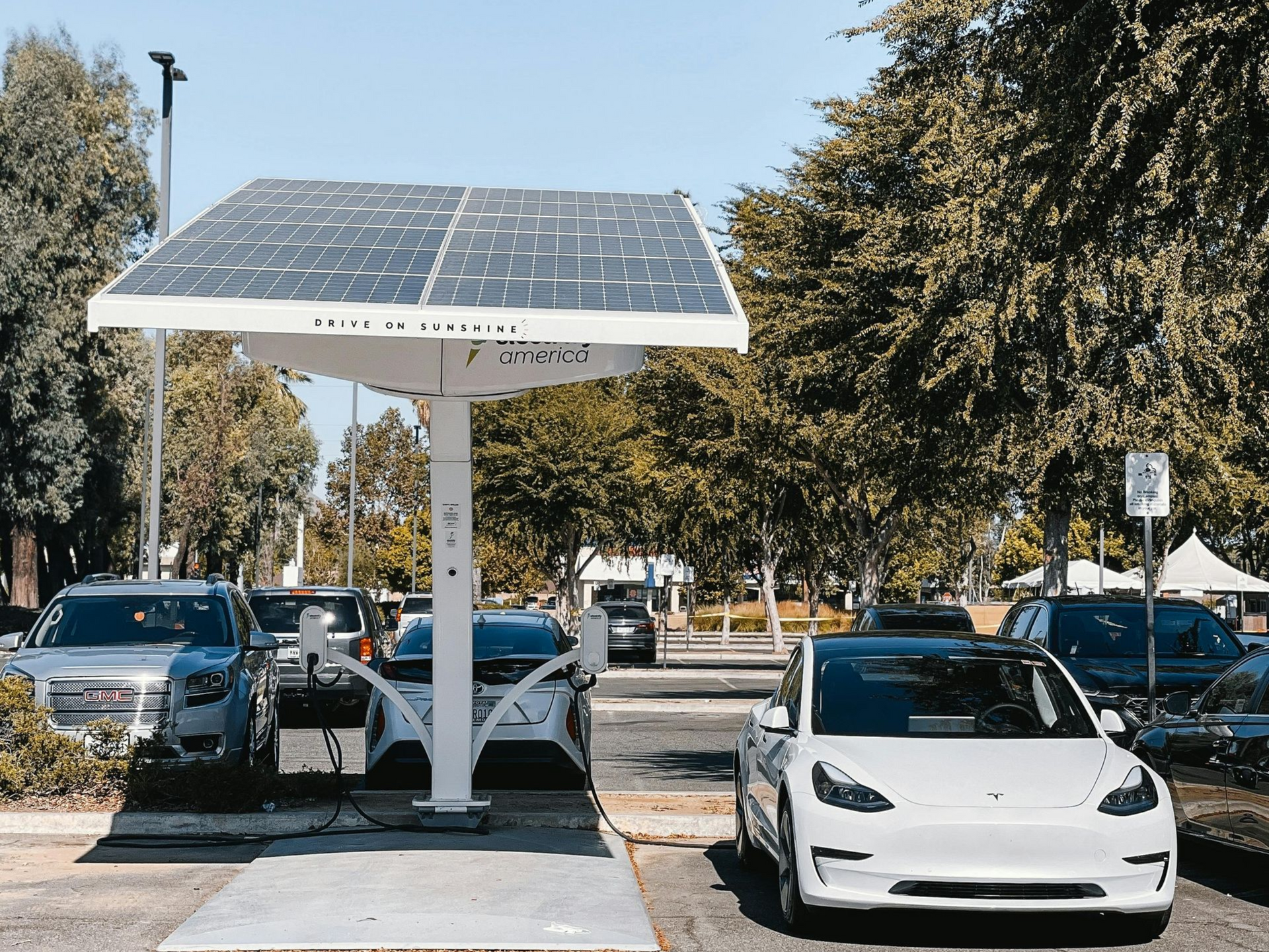 Solar-powered EV charging station with white Tesla. Sunny outdoor setting, cars parked, trees visible.